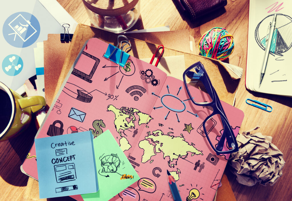 Creative marketing workspace with a tablet showing strategy sketches, world map, icons, sticky notes labeled “Creative Concept” and “Ideas,” glasses, coffee mug, and office supplies on a desk.