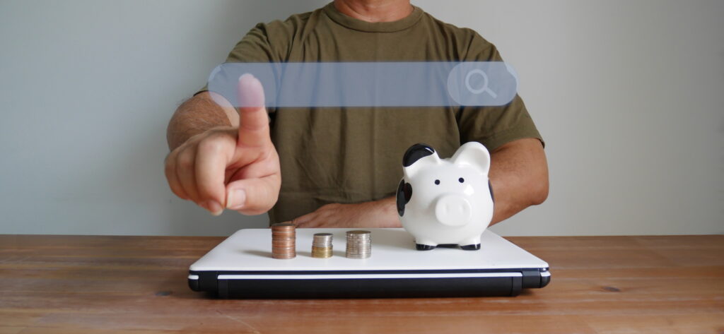 Person pointing at a transparent search bar above a laptop with stacked coins and a piggy bank, symbolizing online savings and financial search tools.