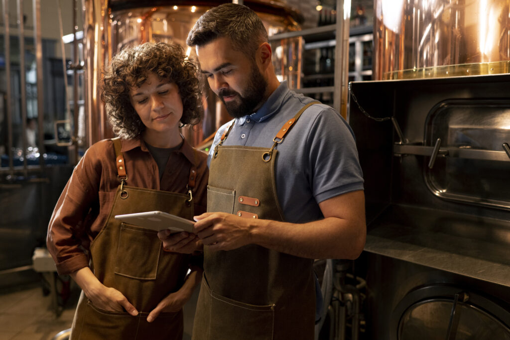 Two brewery owners wearing aprons review information on a tablet inside a working brewery, with brewing tanks and equipment visible in the background.