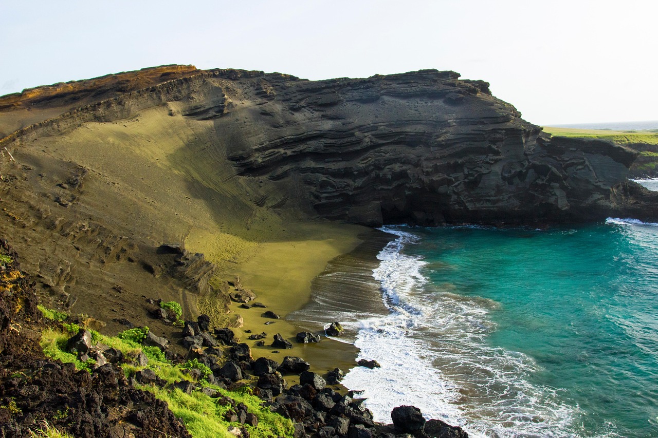 A secluded beach with dramatic volcanic cliffs and greenish sand, where turquoise waves gently meet the rugged shoreline.