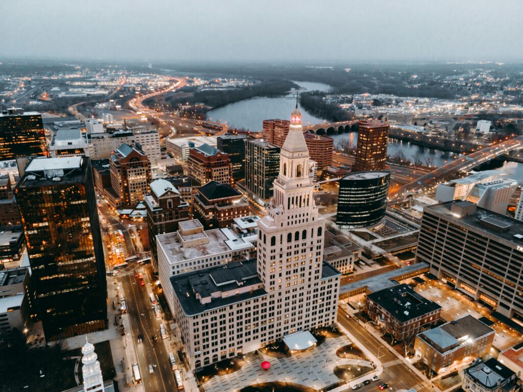 Aerial view of a downtown city at dusk featuring illuminated office buildings, a prominent white clock tower, busy streets with traffic lights, and a river with bridges winding through the background.