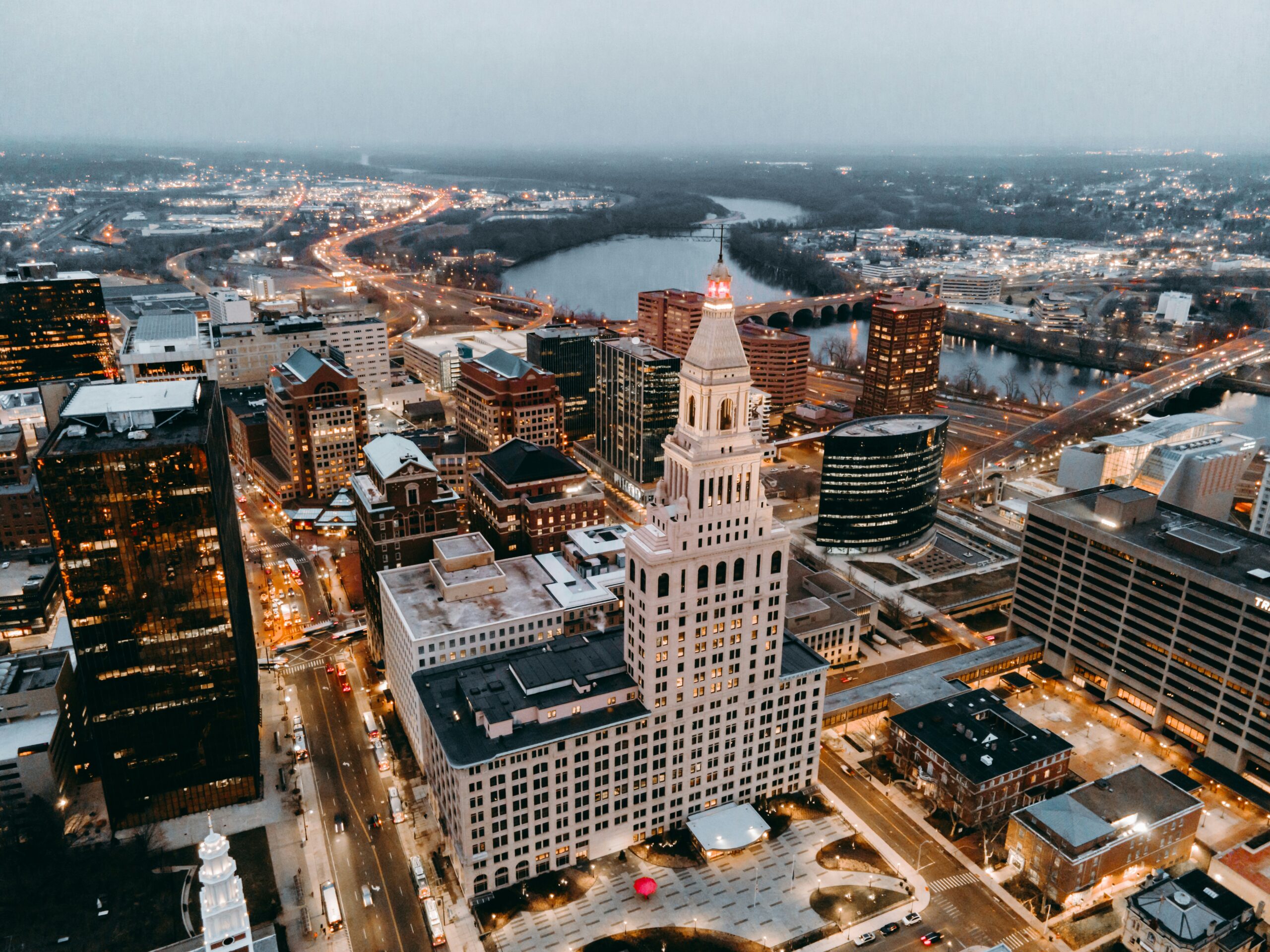 Aerial view of a downtown city at dusk featuring illuminated office buildings, a prominent white clock tower, busy streets with traffic lights, and a river with bridges winding through the background.