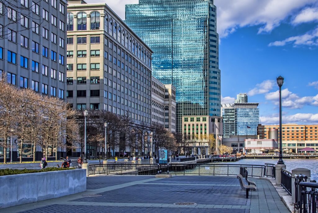 Modern city waterfront with high-rise office buildings, glass skyscraper reflecting blue sky and clouds, pedestrian walkway with benches and railing along the water, and people strolling on a clear day.