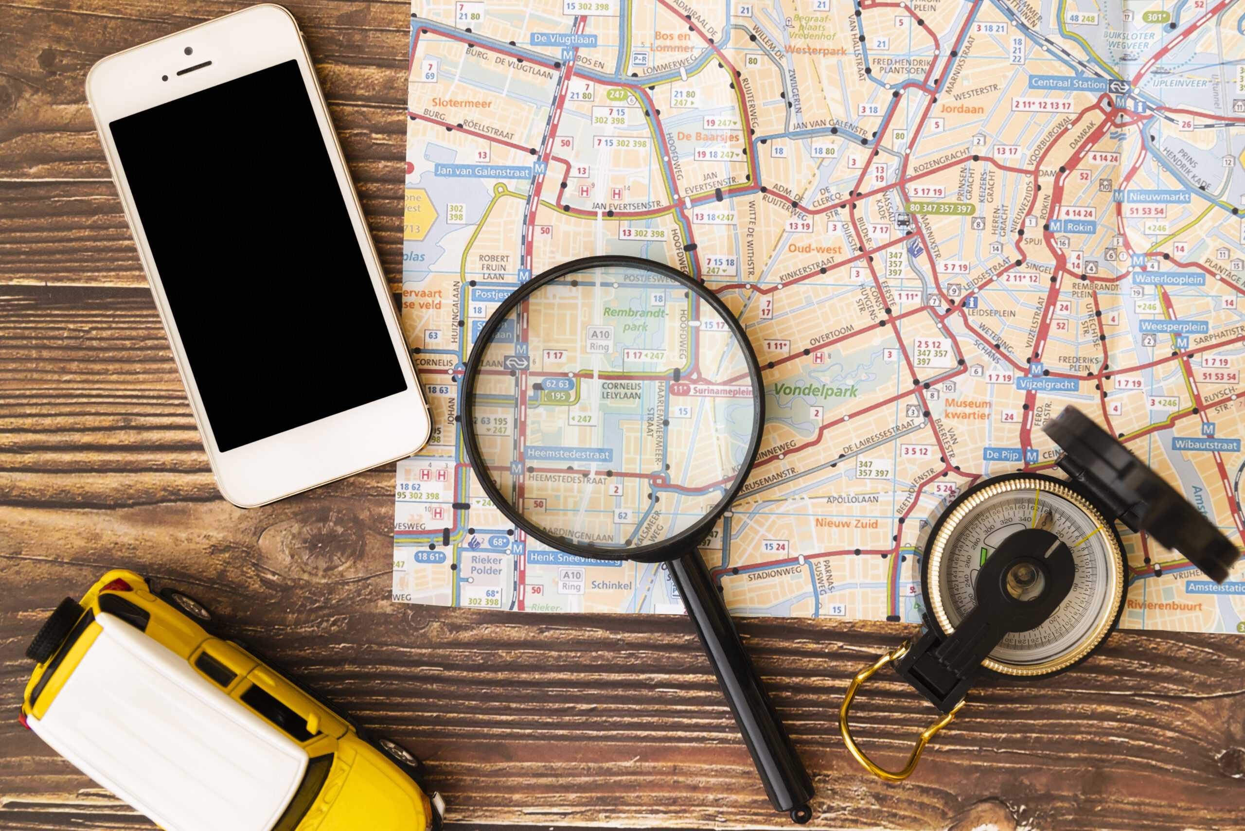 Top view of a wooden table with a city map spread out, a magnifying glass centered over the map, a smartphone with a blank screen, a compass, and a small yellow toy van, suggesting navigation and travel planning.