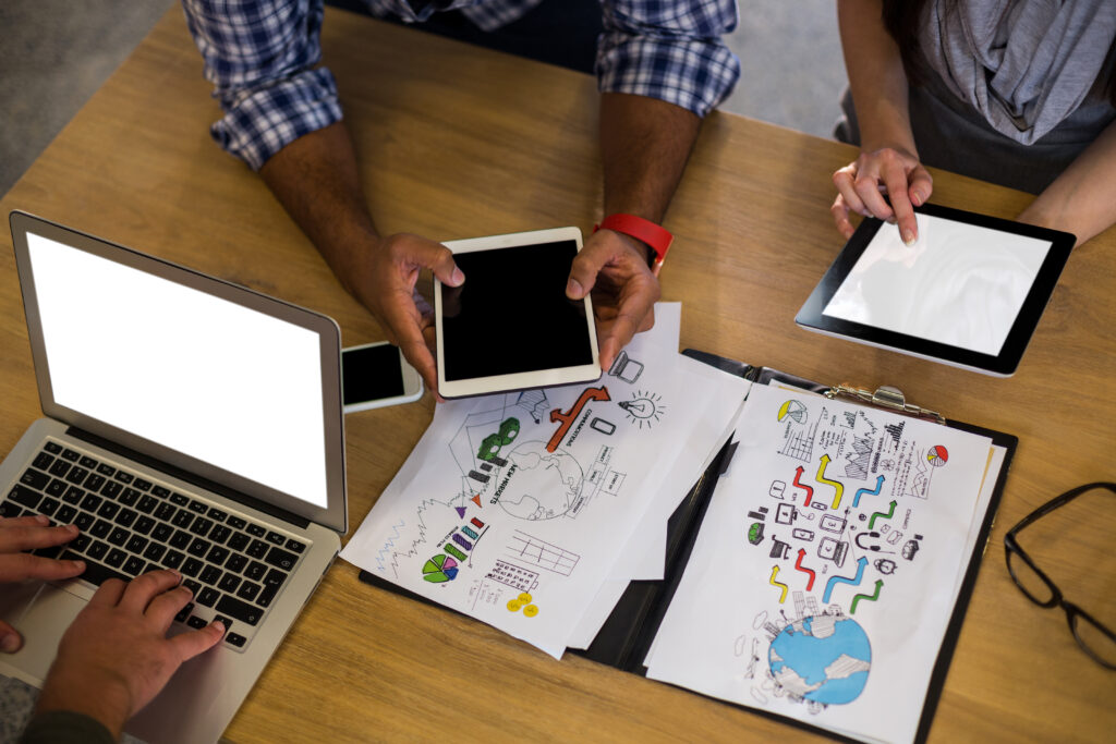 Overhead view of a team meeting at a wooden table with a laptop, tablets, and printed charts showing colorful graphs and diagrams, as two people hold tablets and collaborate on a digital marketing or business strategy plan.