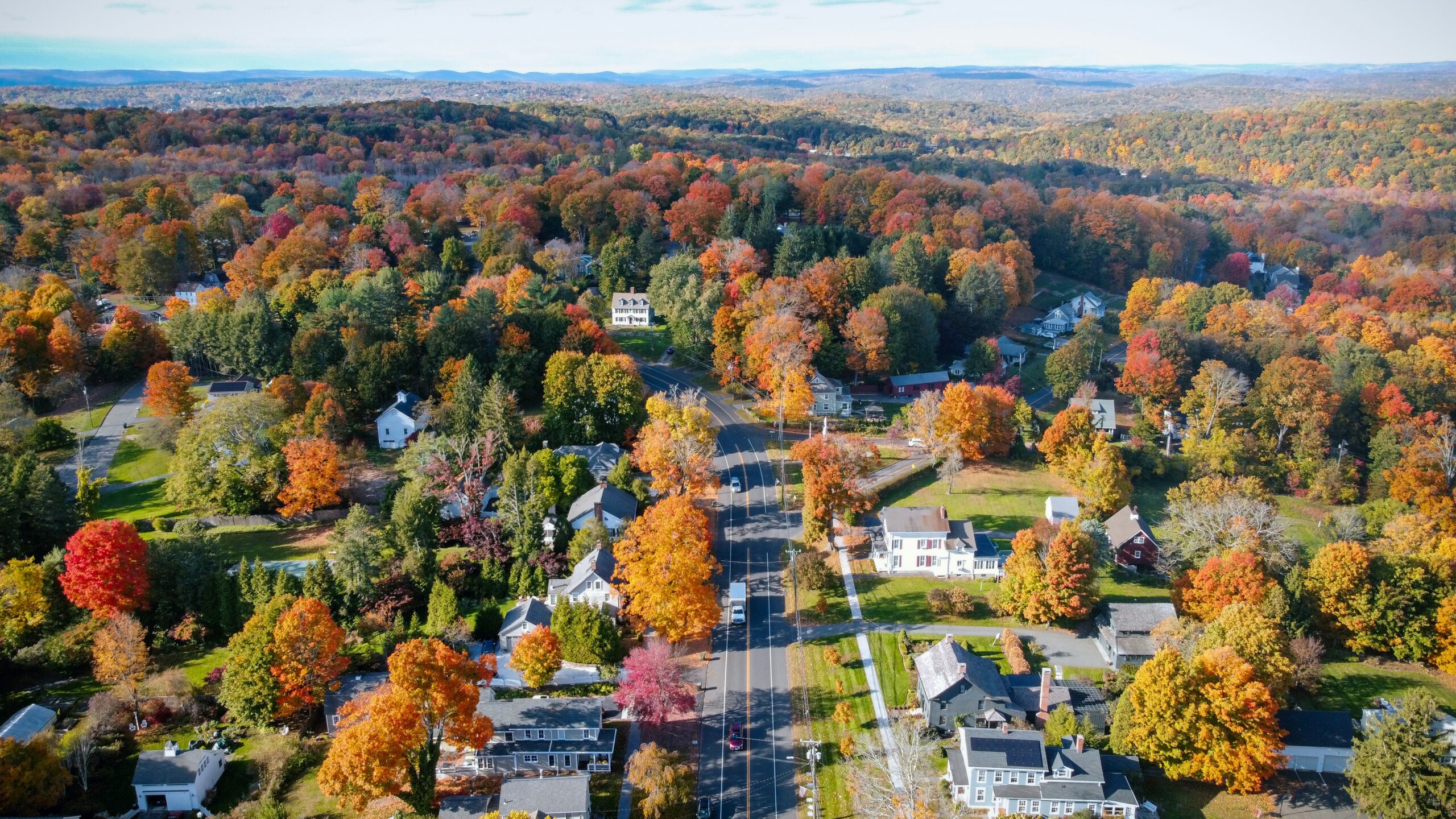 Aerial view of a Connecticut residential town in autumn, showing tree-lined neighborhoods with colorful fall foliage, houses, and a main road running through the community.