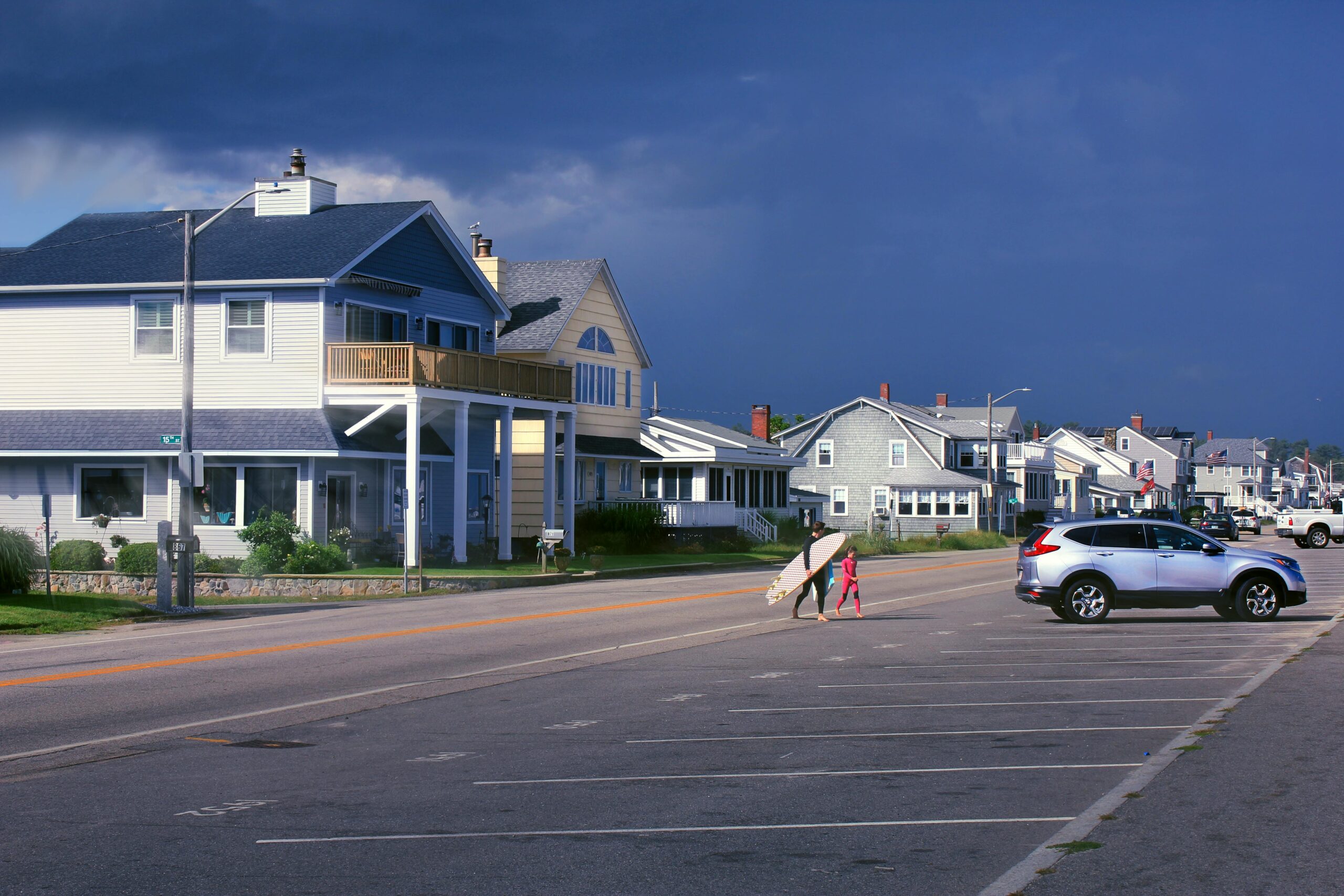 A coastal street with beach houses under dark storm clouds. A man carrying a surfboard walks with a child across the road near a parked SUV.