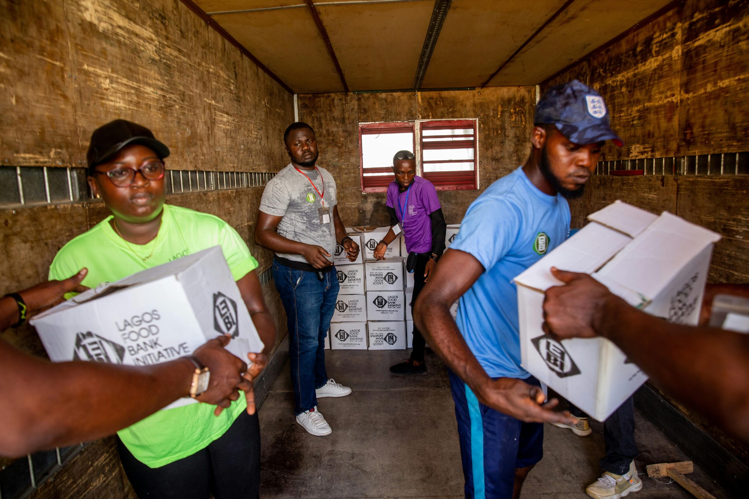 Volunteers inside the back of a truck unloading and handing out labeled food aid boxes, with stacks of cartons in the background and people working together in a confined cargo space.