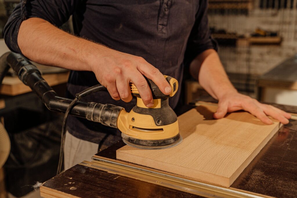 Close-up of a person using an electric orbital sander on a wooden board in a workshop, smoothing the surface while holding the wood steady on a workbench.