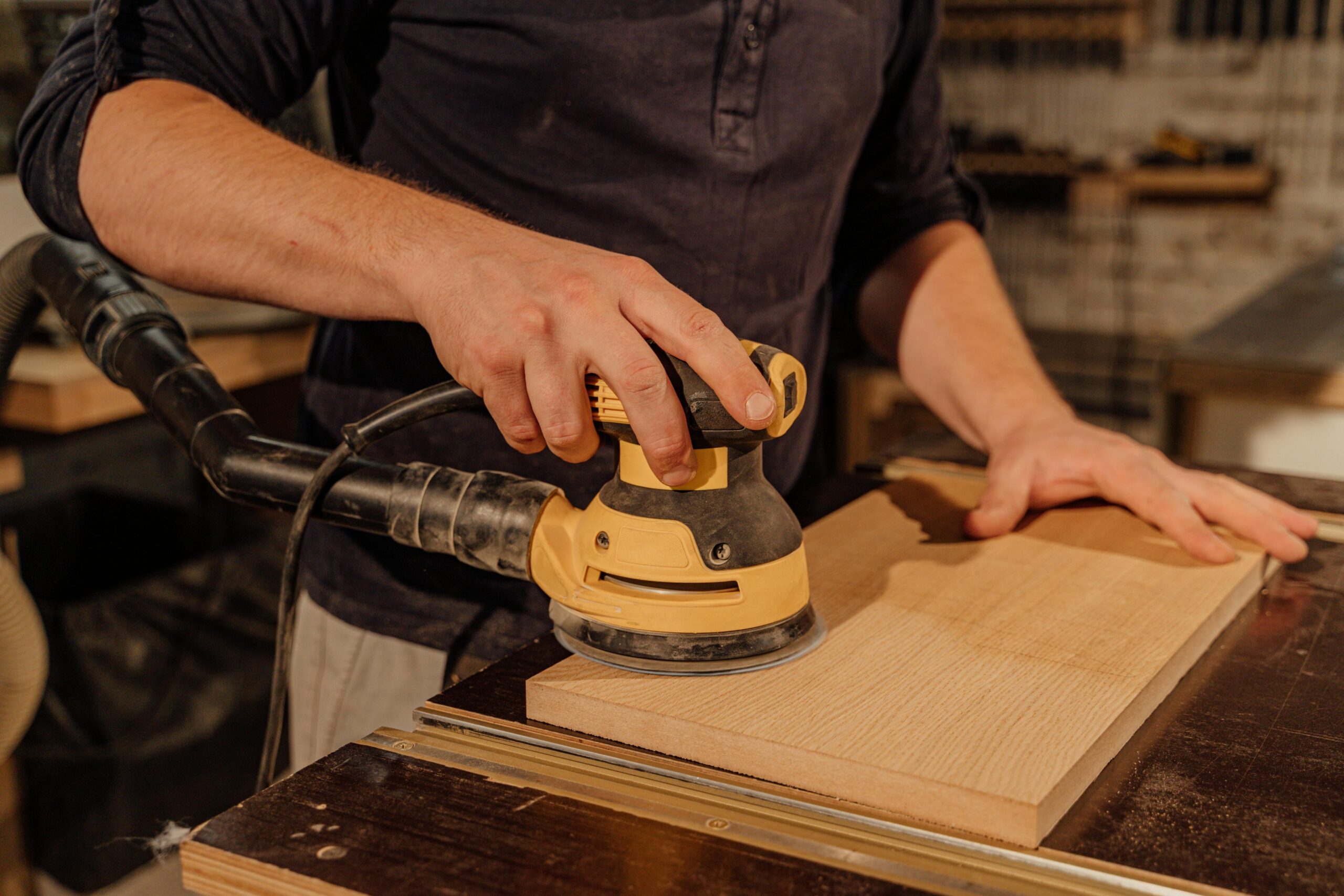 Close-up of a person using an electric orbital sander on a wooden board in a workshop, smoothing the surface while holding the wood steady on a workbench.