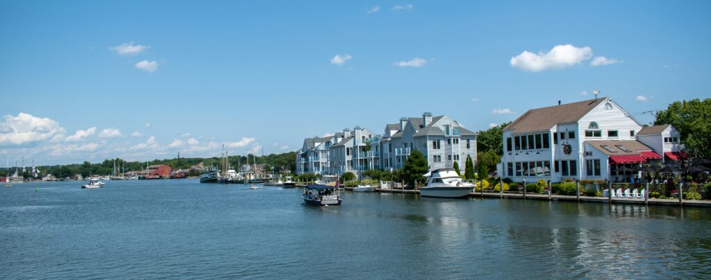 Waterfront homes and boats line a calm harbor under a clear blue sky, with docks and a seaside restaurant along the shore.