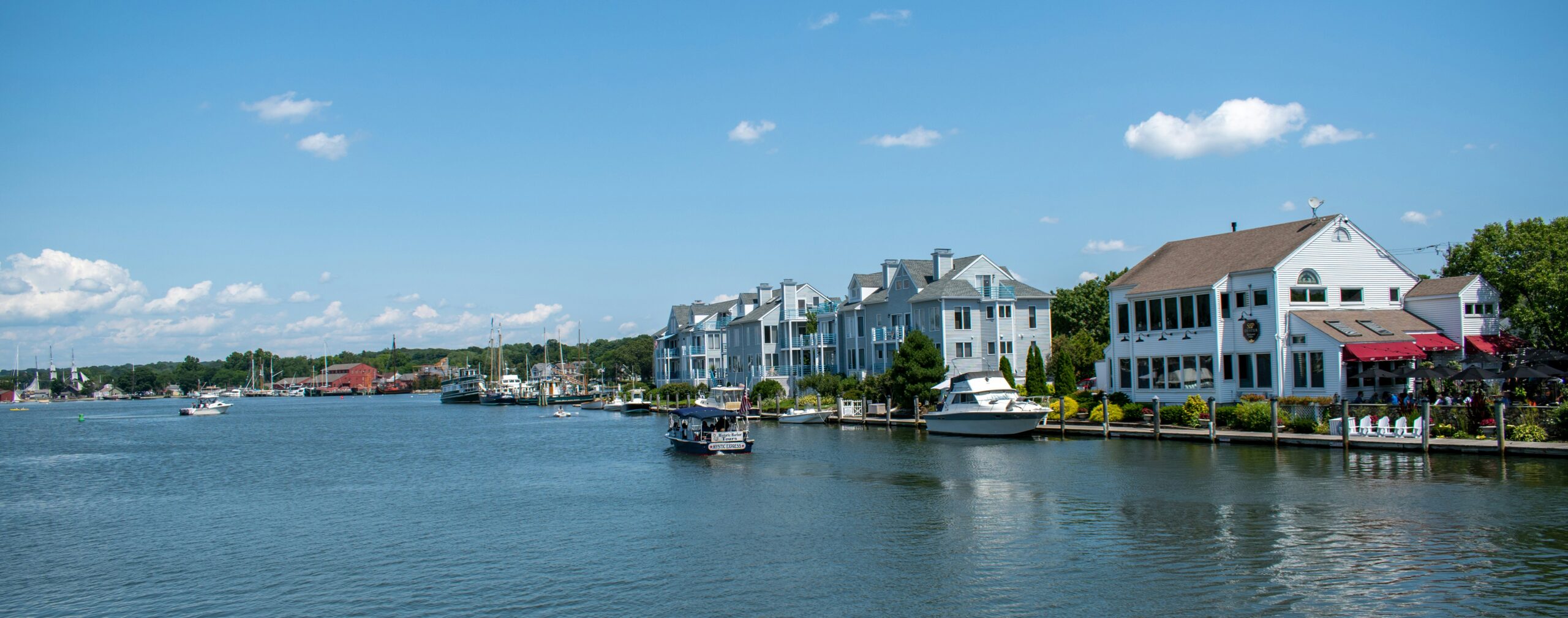 Waterfront homes and boats line a calm harbor under a clear blue sky, with docks and a seaside restaurant along the shore.