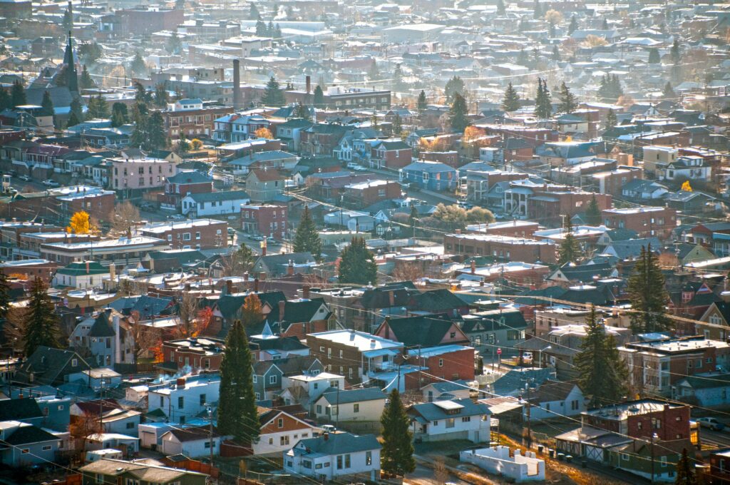 Aerial view of a Montana town with closely packed homes, brick buildings, and tree-lined streets, set against a lightly misty backdrop with autumn foliage scattered throughout the neighborhood.