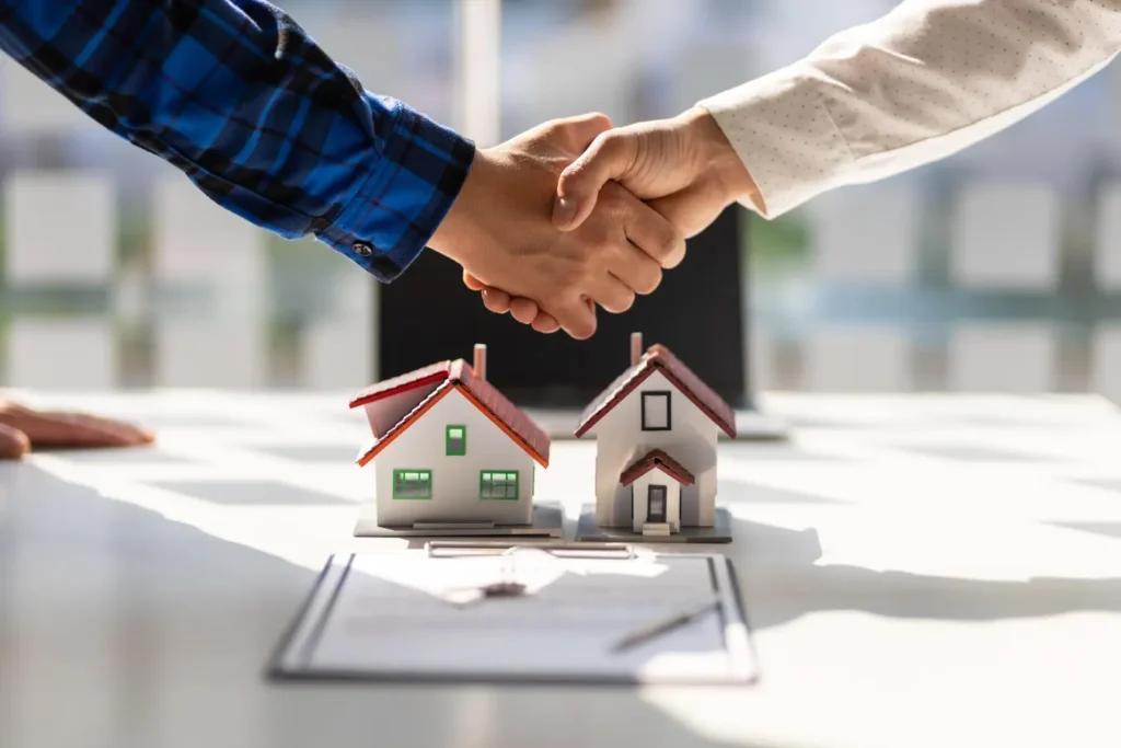 Real estate agents shaking hands over a table with miniature model houses and paperwork, symbolizing a successful home purchase or property agreement.