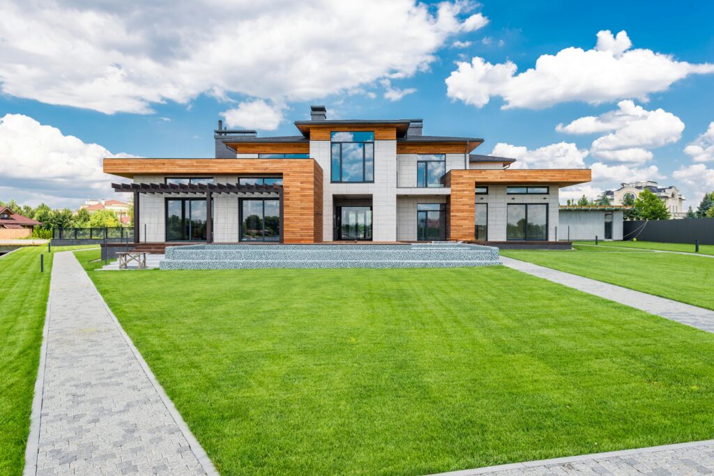 Modern two-story luxury home with a minimalist design, featuring wood and stone exterior, large floor-to-ceiling windows, a manicured green lawn, and paved walkways under a bright blue sky with scattered clouds.