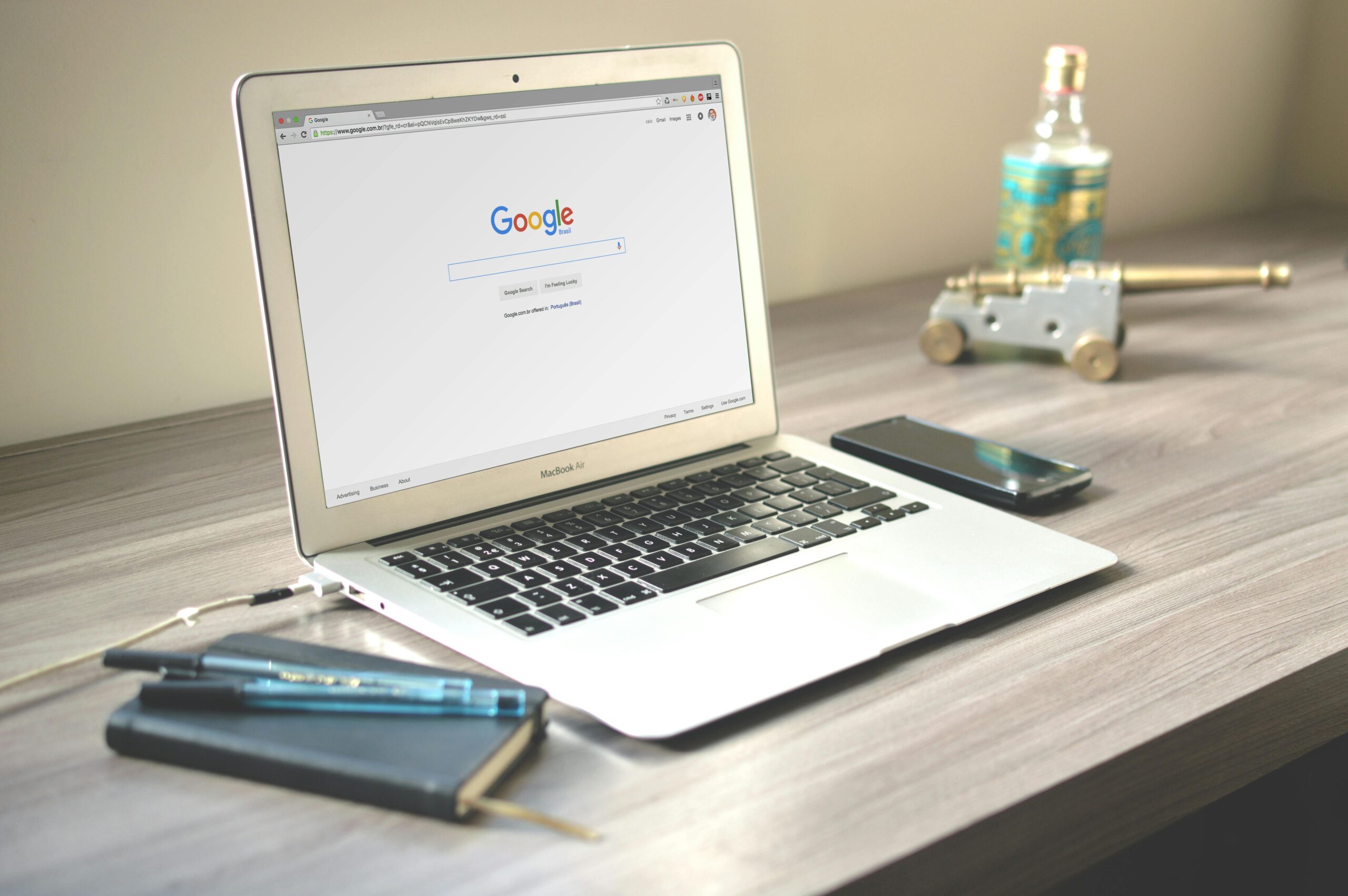 MacBook Air laptop displaying the Google homepage on a desk with a smartphone, notebook, and pen nearby.