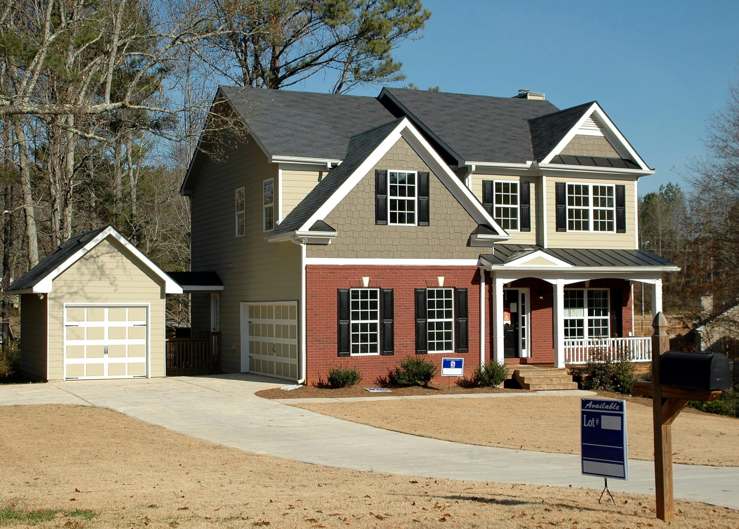Two-story suburban house with beige siding and red brick exterior, black shutters, a covered front porch with white columns, and a detached garage connected by a driveway. A “lot available” sign and mailbox sit near the front yard.