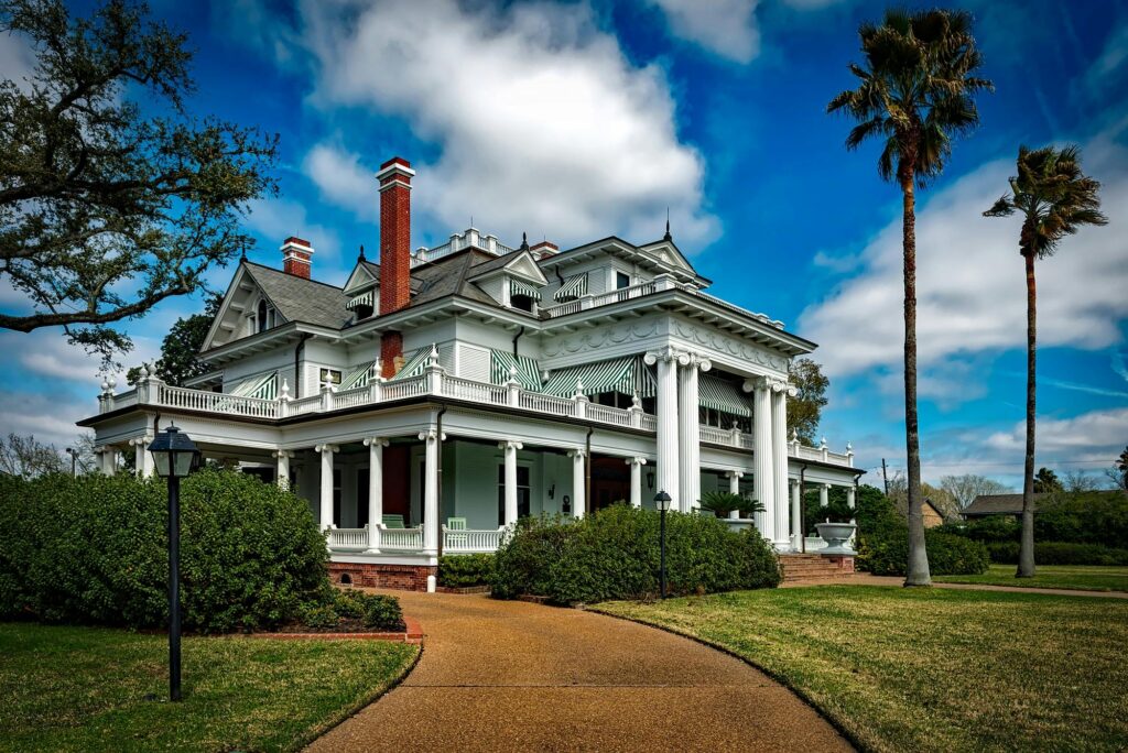 Large historic mansion with white columns, wraparound porch, decorative railings, and green-striped awnings, surrounded by palm trees, landscaped bushes, and a curved walkway under a bright blue sky.