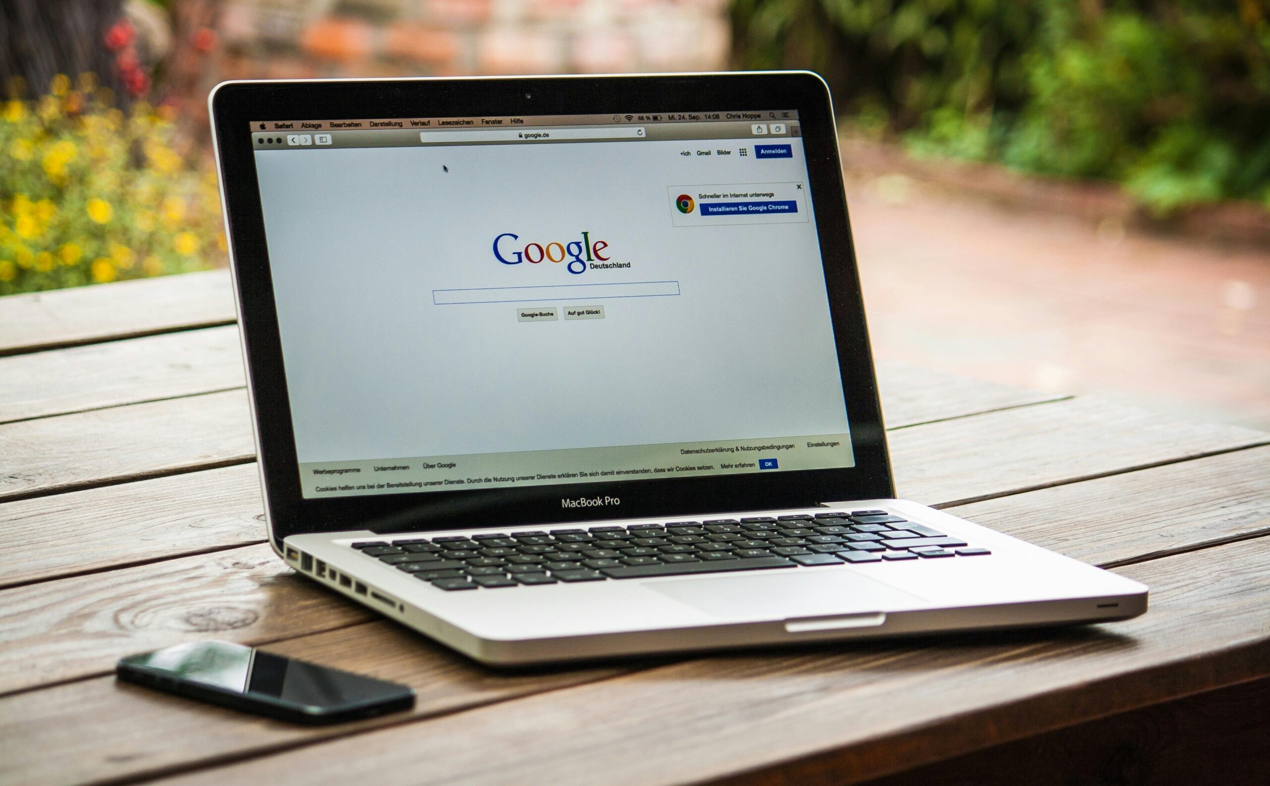 Laptop on a wooden outdoor table displaying the Google search homepage, with a smartphone lying beside it.