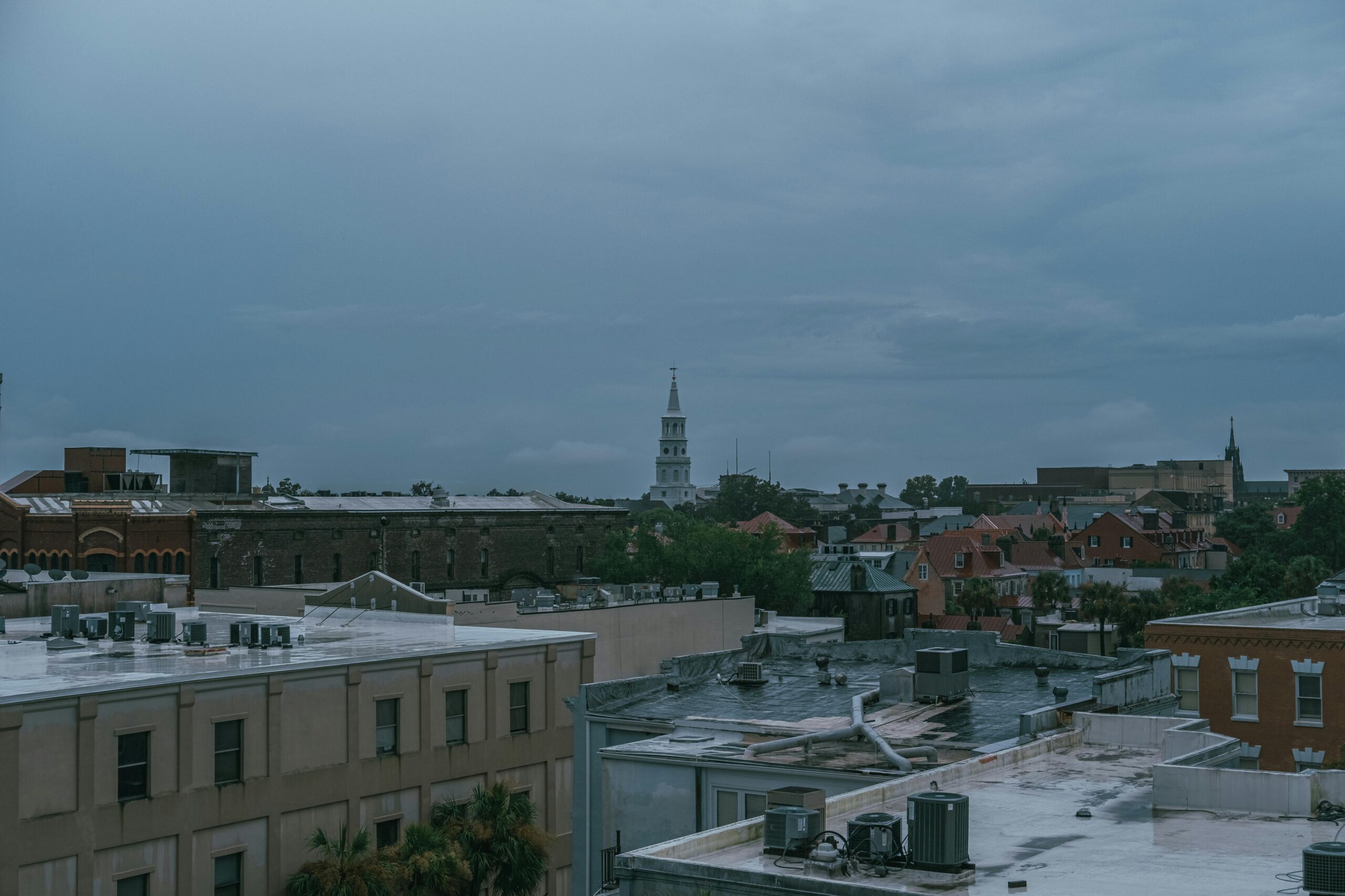 Rooftop view of a historic downtown cityscape under overcast skies, with a white church steeple rising above surrounding brick buildings and green trees.