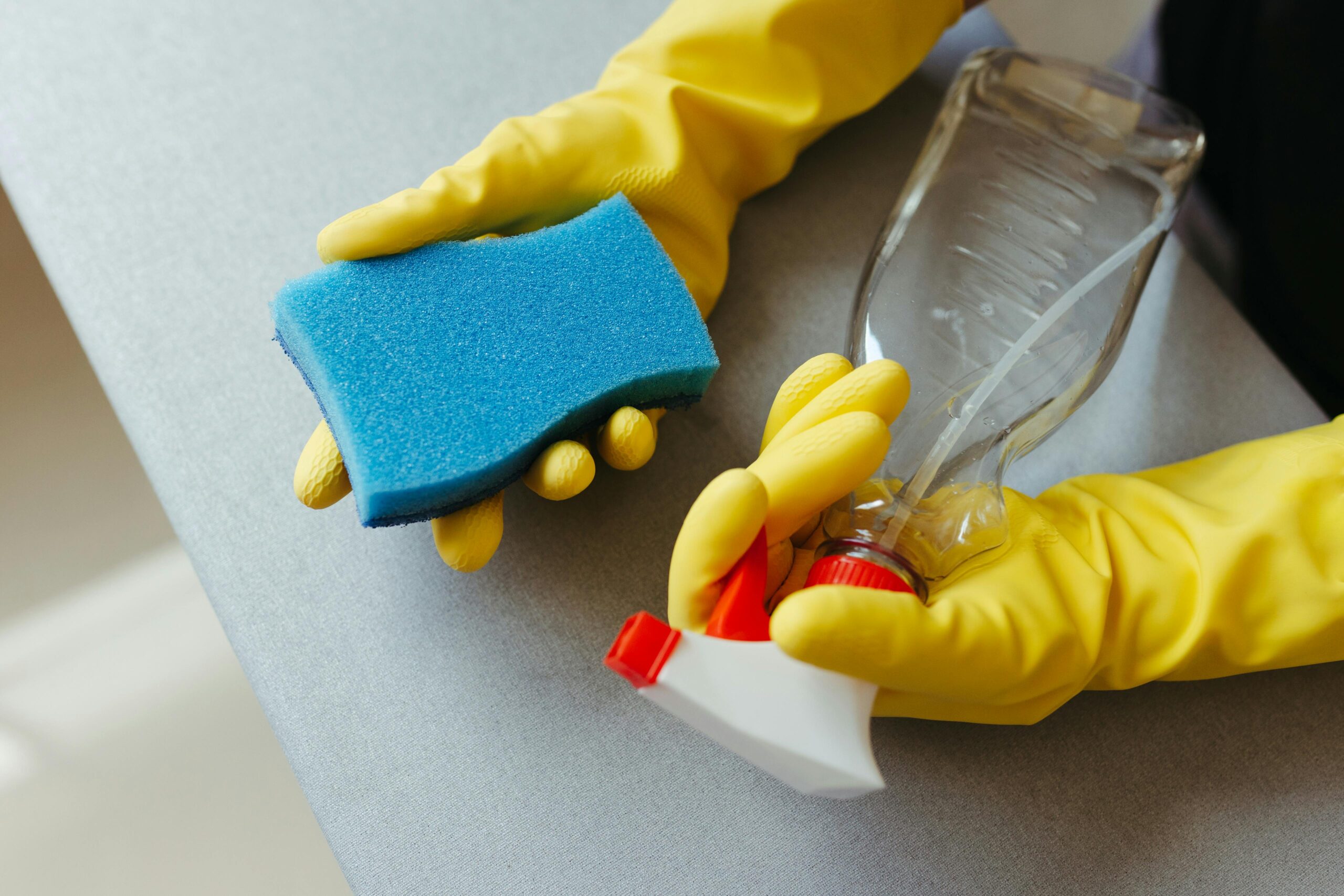 Hands wearing yellow rubber gloves holding a blue sponge and a spray bottle while cleaning a gray surface.