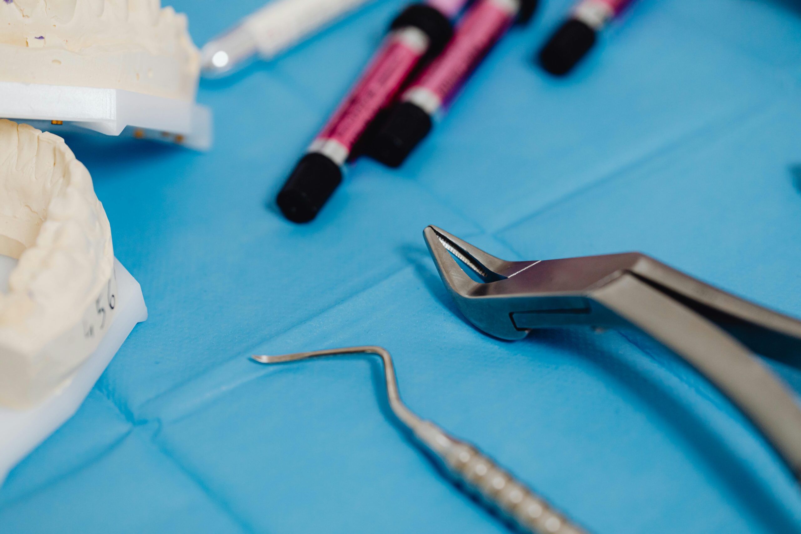 Dental instruments including extraction forceps, a scaler, composite syringes, and a plaster tooth model arranged on a blue surface.