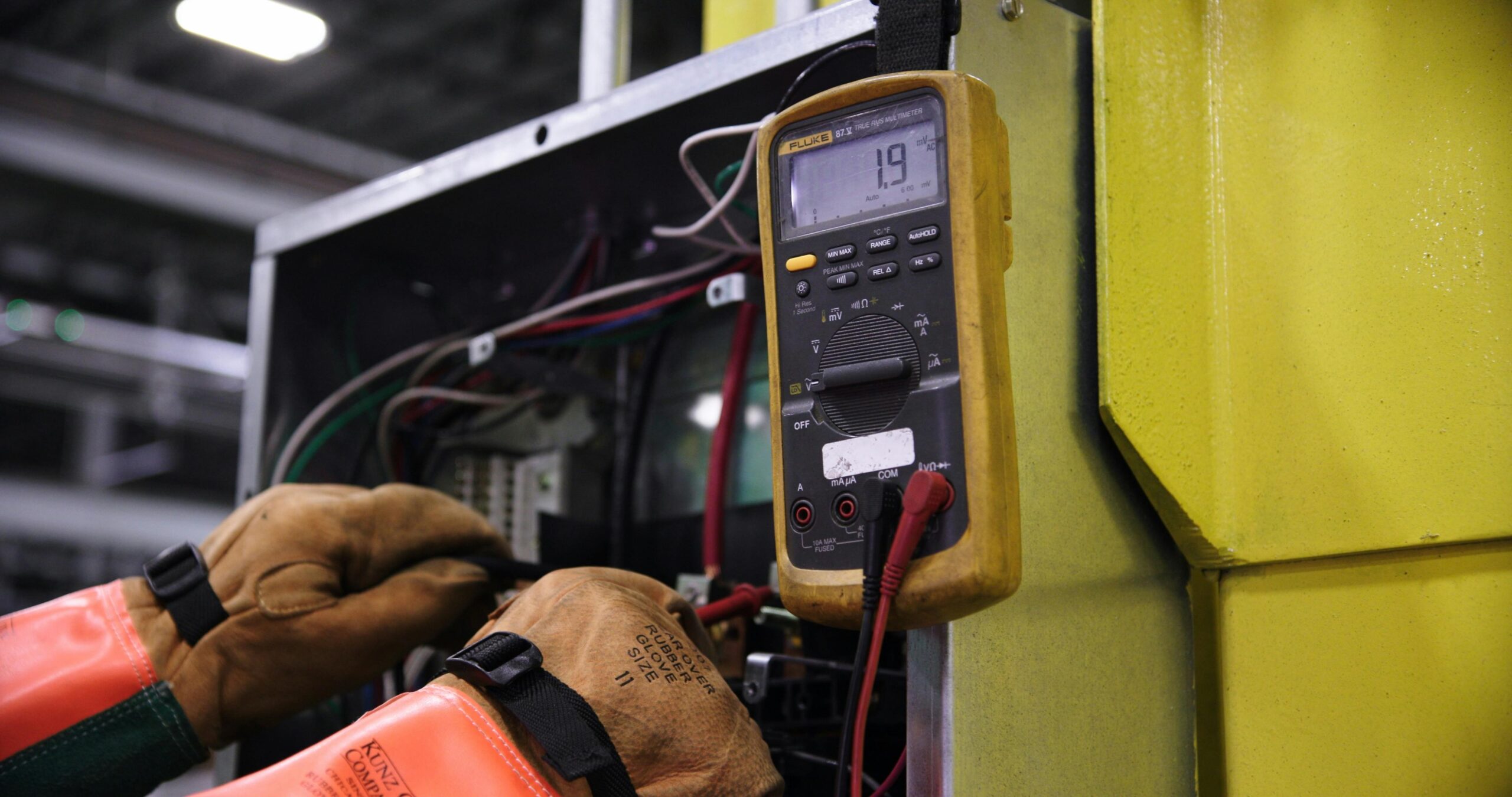 A worker in rubber safety gloves and an orange jacket using a Fluke 87V multimeter to test industrial electrical wiring inside a control panel.