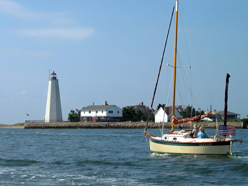 Sailboat on open water near a coastal lighthouse, with houses along the shoreline under a clear blue sky.