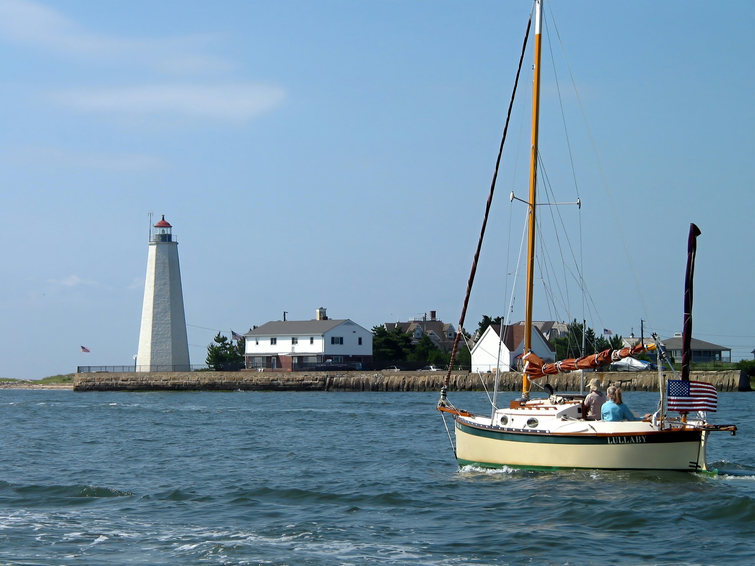 Sailboat on open water near a coastal lighthouse, with houses along the shoreline under a clear blue sky.