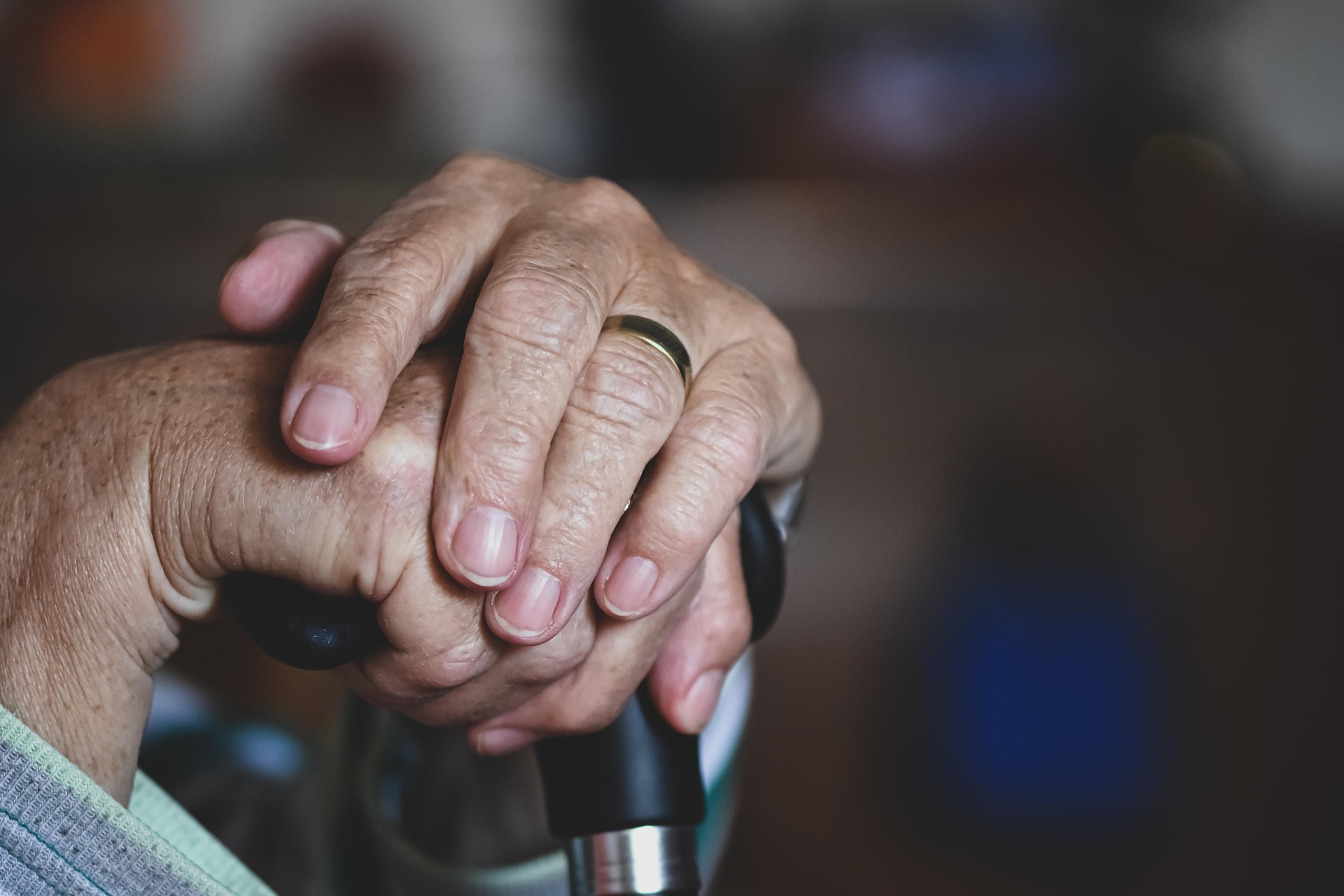 Close-up of an elderly person's hands resting on a cane, showing aging skin and a wedding band in a softly lit indoor setting.