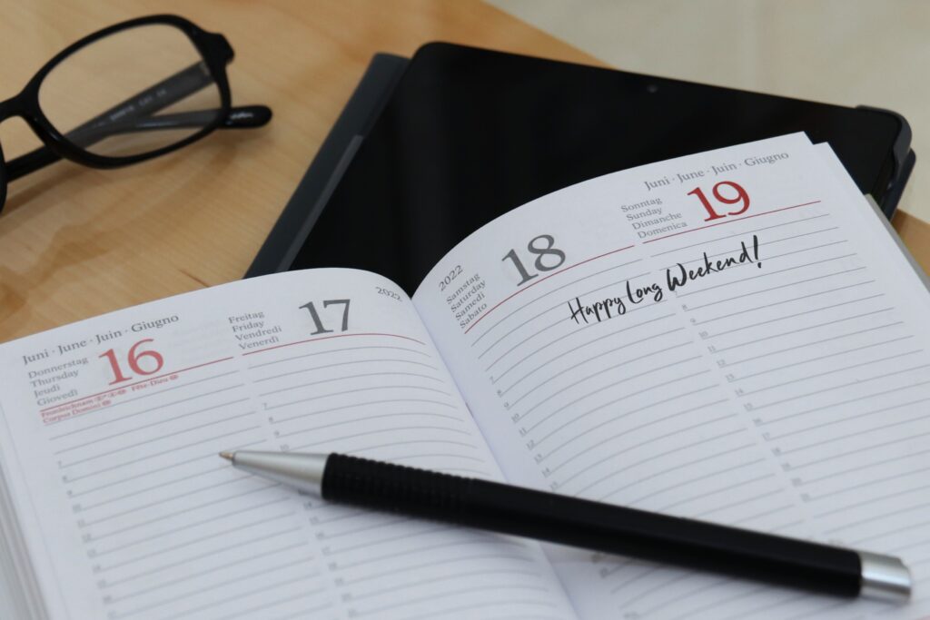 Open planner on a desk showing June 16–19 with “Happy Long Weekend!” written, a pen resting across the pages, and glasses nearby.