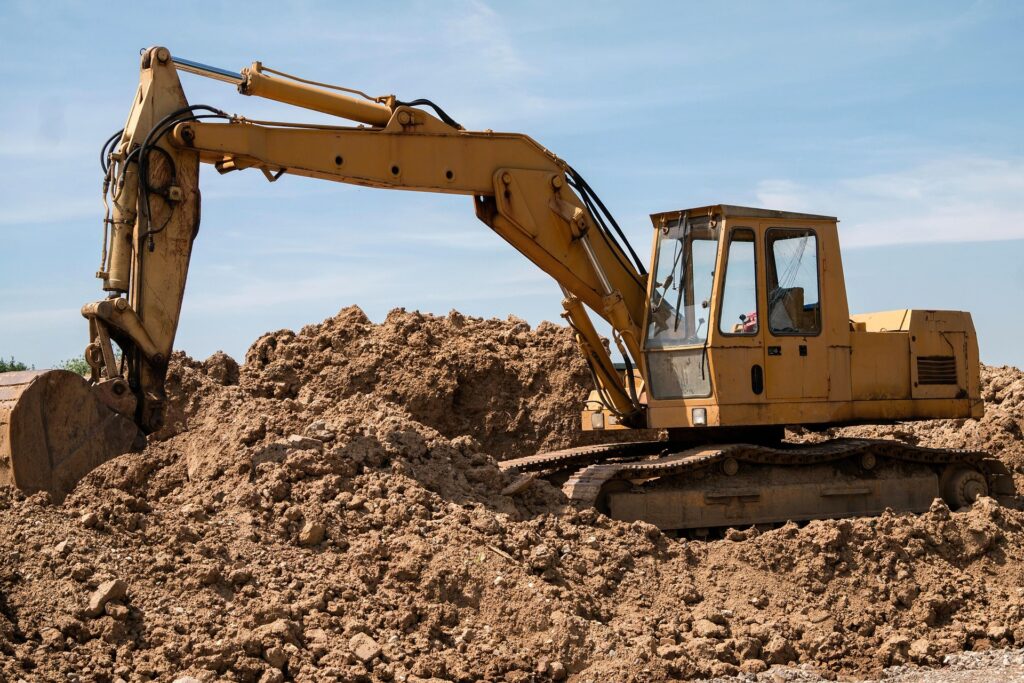 Yellow excavator digging through large piles of dirt at an outdoor construction site under a clear blue sky.
