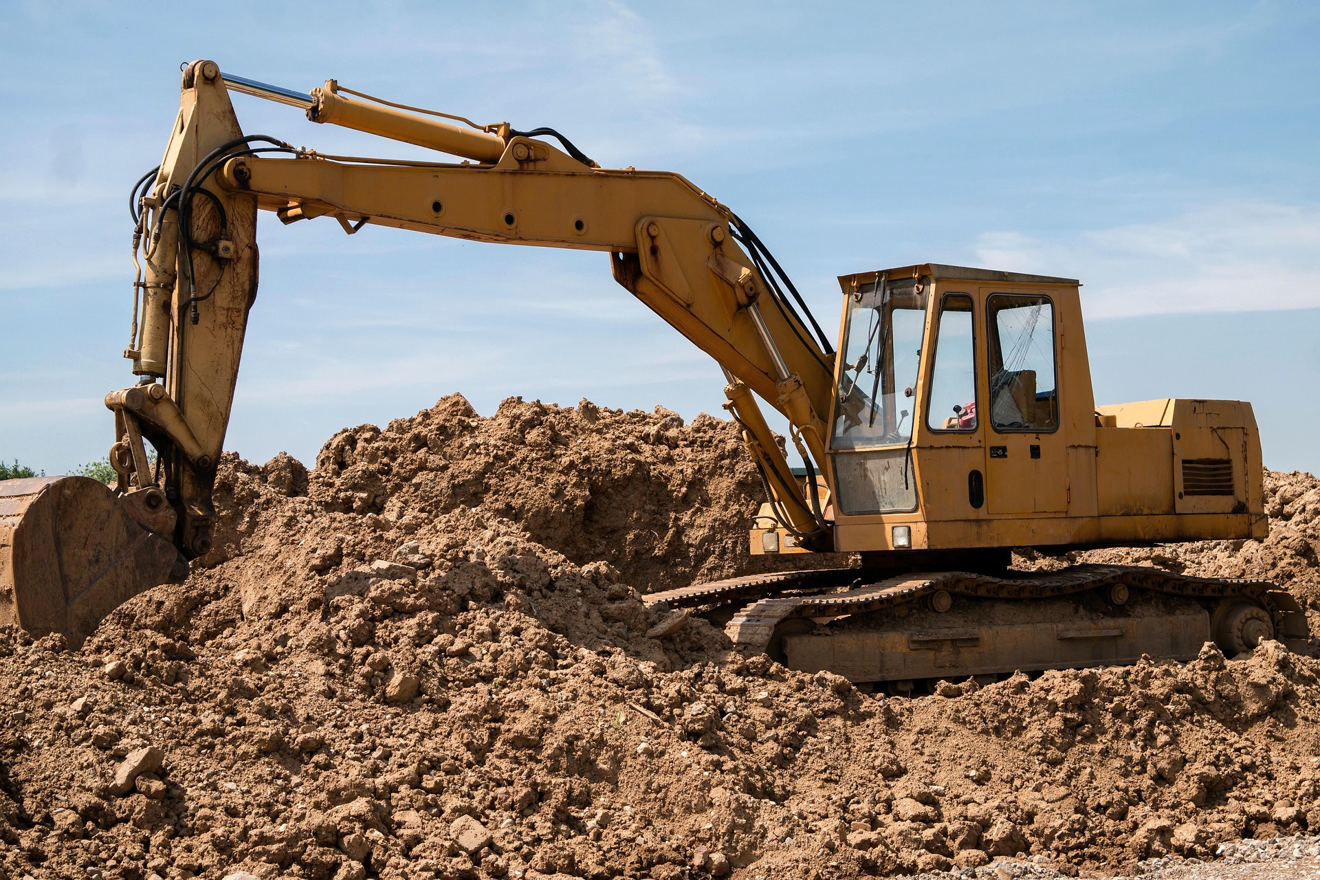 Yellow excavator digging through large piles of dirt at an outdoor construction site under a clear blue sky.