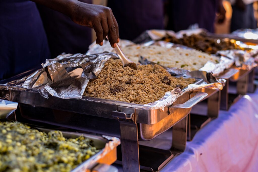 Buffet-style catering setup with trays of seasoned rice, vegetables, and hot dishes being served at an indoor event.