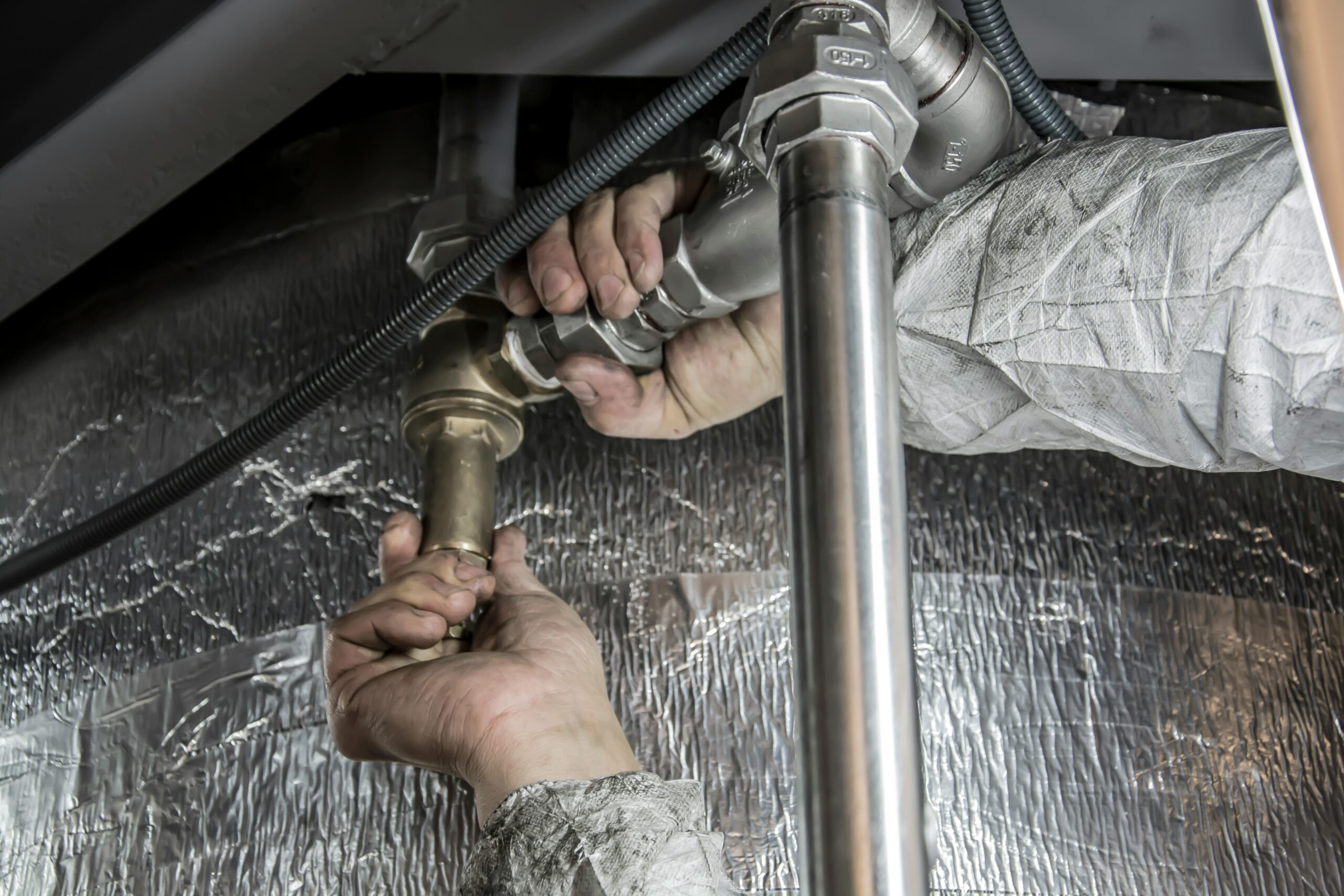 Close-up of a technician tightening industrial metal piping and valves while working on insulated mechanical equipment.