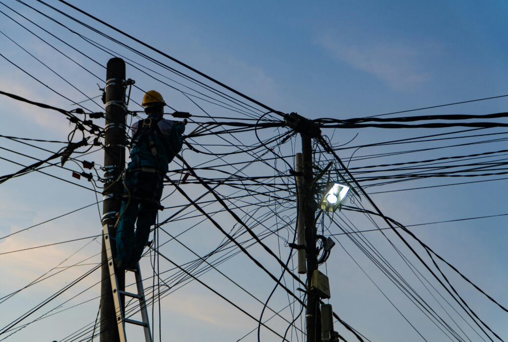 Utility worker climbing a ladder on a power pole surrounded by tangled overhead electrical and communication wires at dusk.