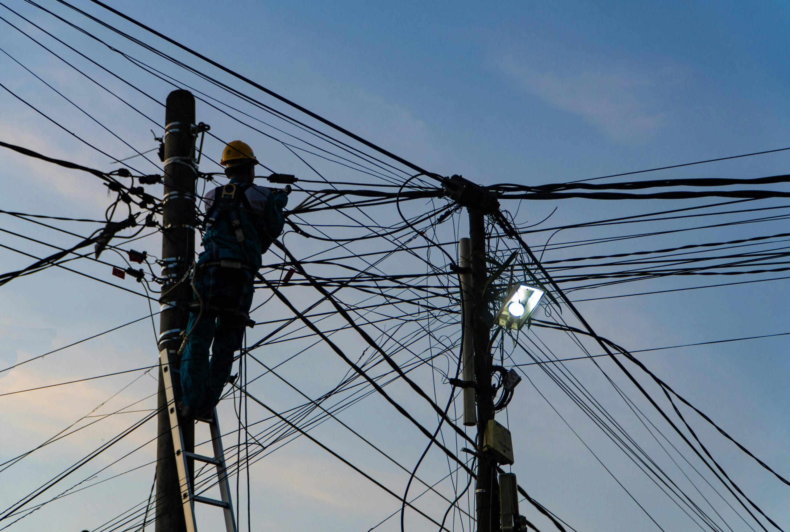 Utility worker climbing a ladder on a power pole surrounded by tangled overhead electrical and communication wires at dusk.