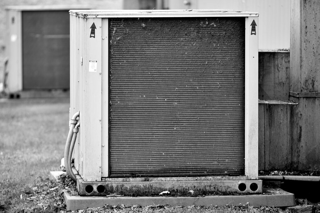 Black-and-white photo of an outdoor commercial HVAC condenser unit with visible metal fins and utility connections beside a building.