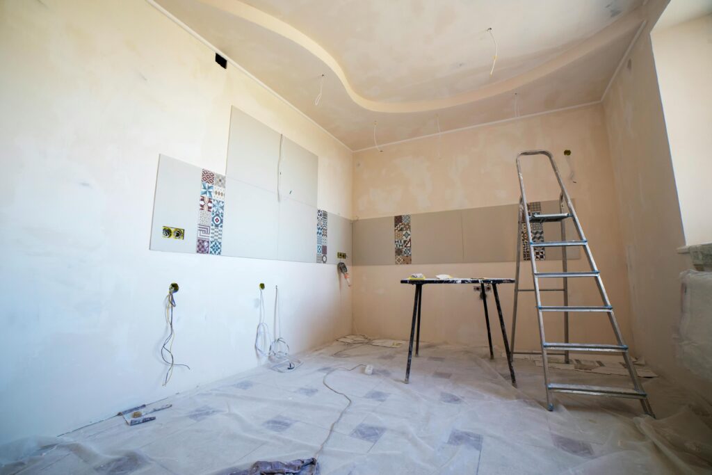 Unfinished room under renovation with partially tiled walls, exposed wiring, a ladder, and a work table, with protective covering on the floor.
