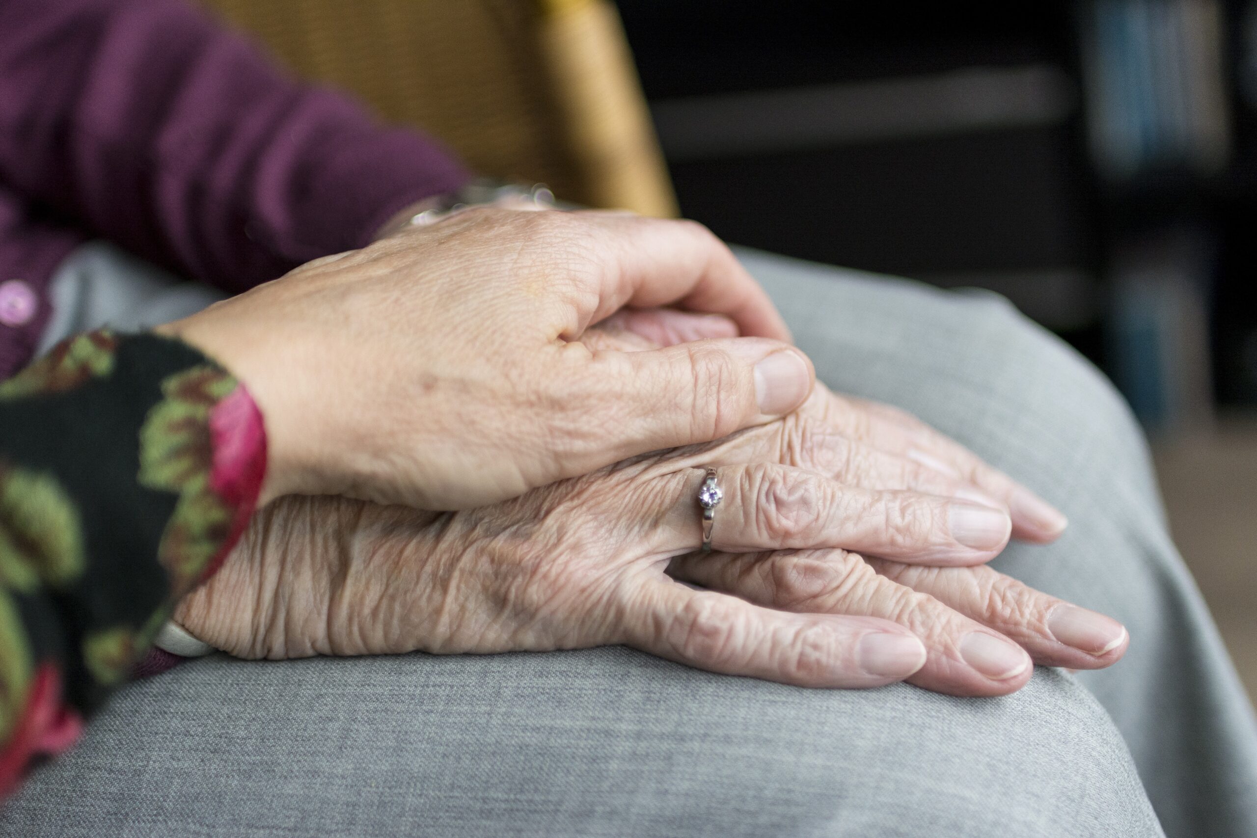 Close-up of one person gently holding another older person’s hand, showing care, support, and compassion, with a ring visible on the older hand.