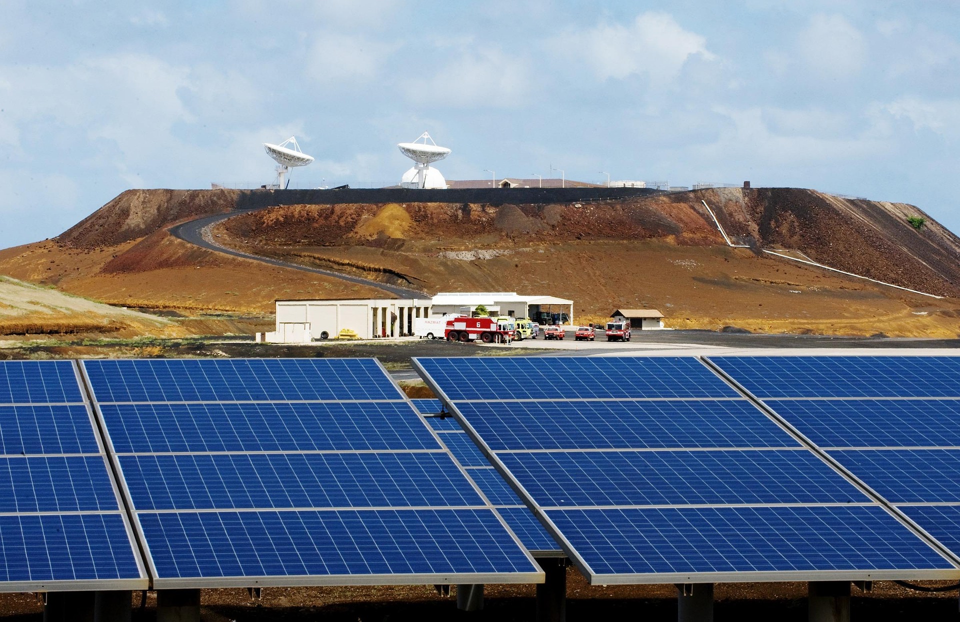 Solar panels installed in the foreground of a remote energy facility, with satellite dishes and support buildings on a rocky hillside in the background.