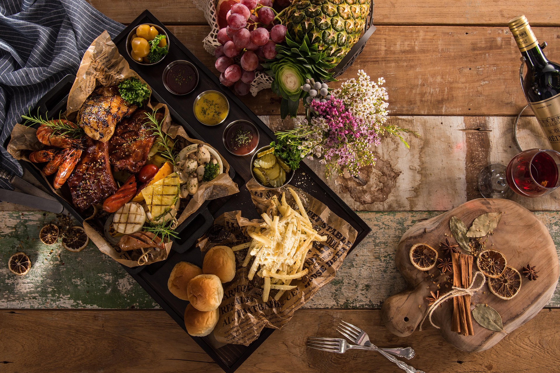 Rustic feast platter with grilled meats, shrimp, vegetables, fries, bread rolls, fruit, sauces, and red wine on a wooden table.