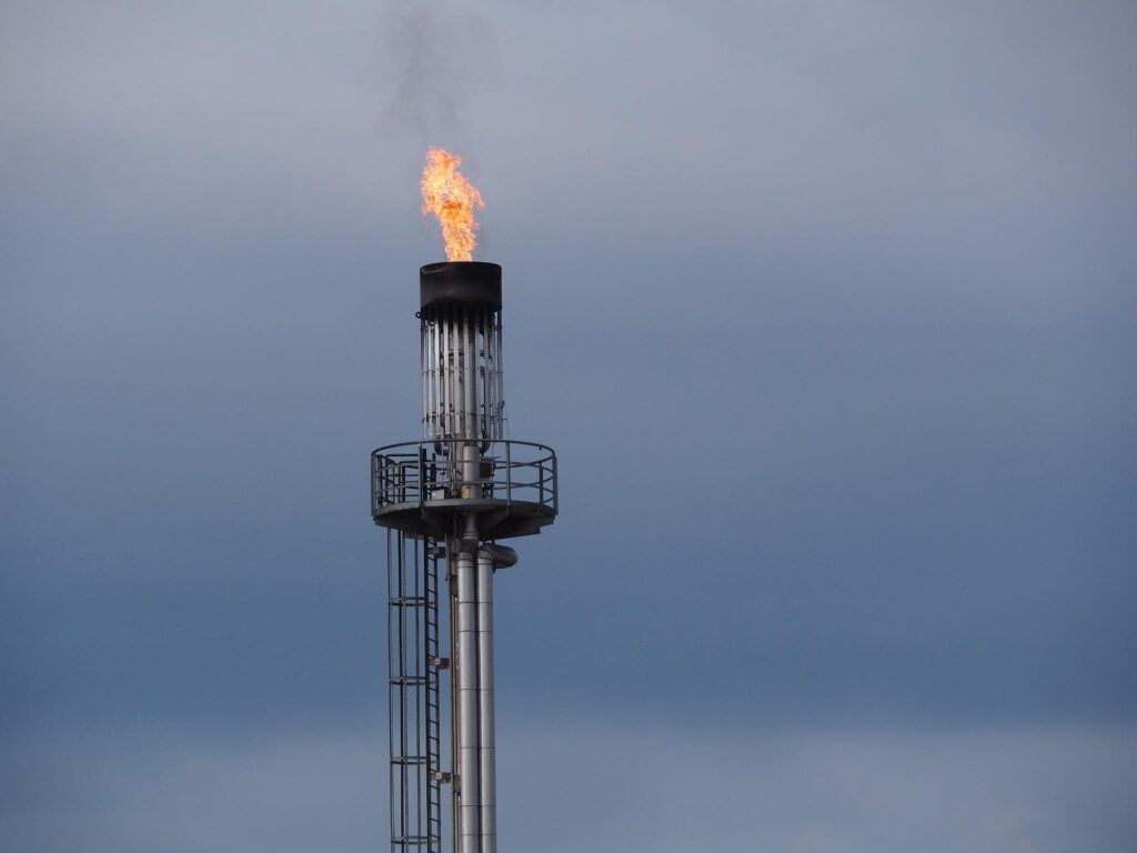 Industrial flare stack with a visible flame burning at the top against a cloudy sky.