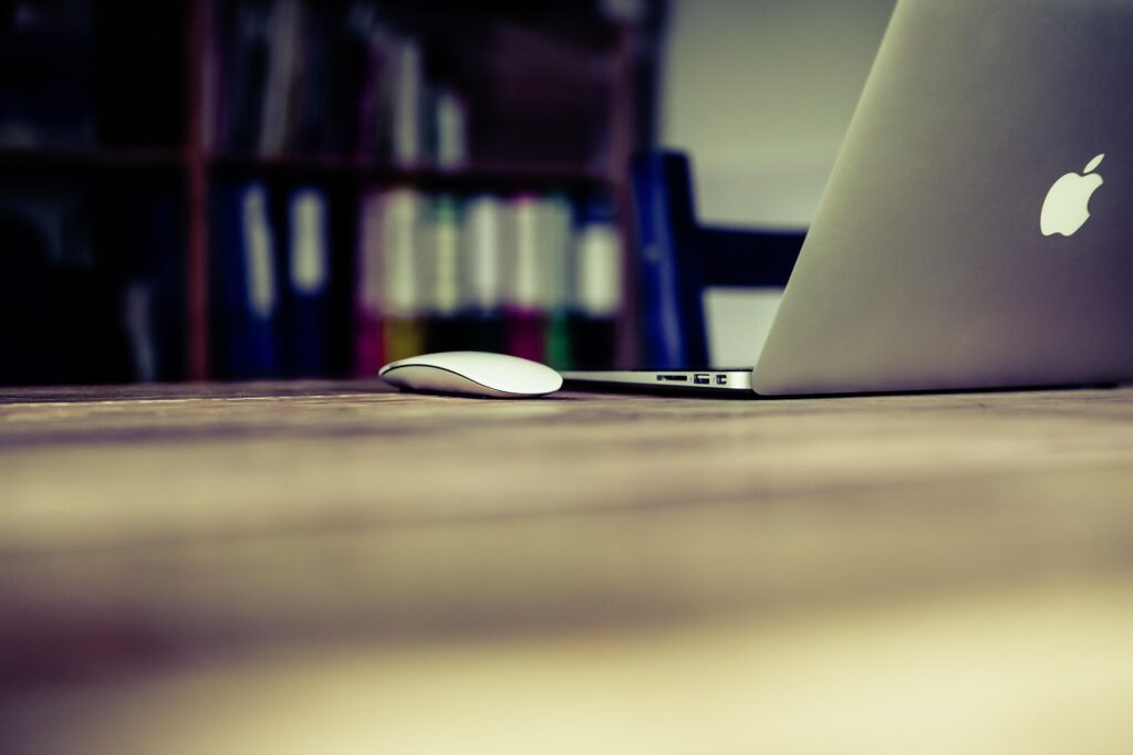 Silver laptop and wireless mouse on a wooden desk with blurred bookshelves in the background.