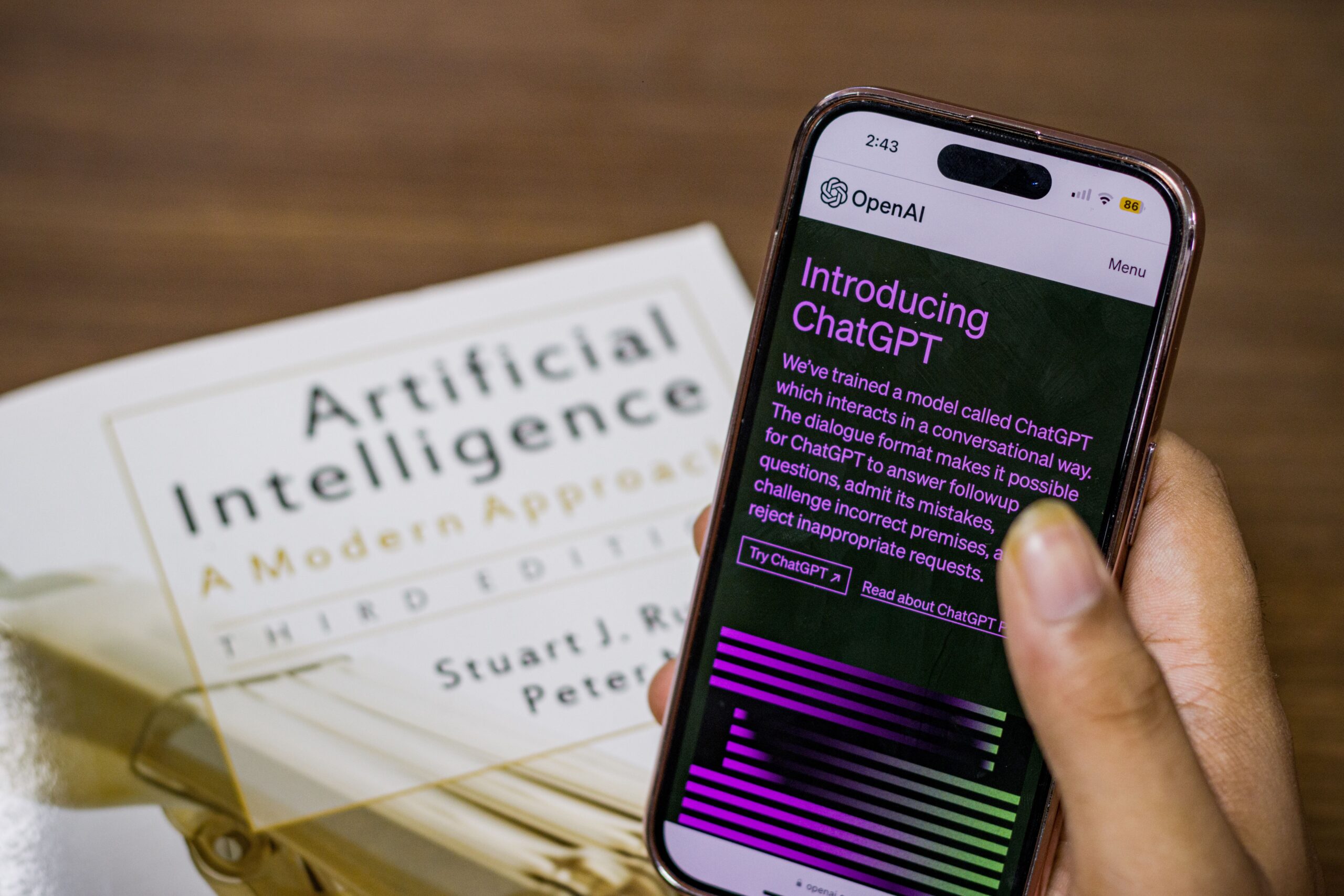 Person holding a smartphone displaying the ChatGPT introduction page, with an artificial intelligence book blurred in the background.
