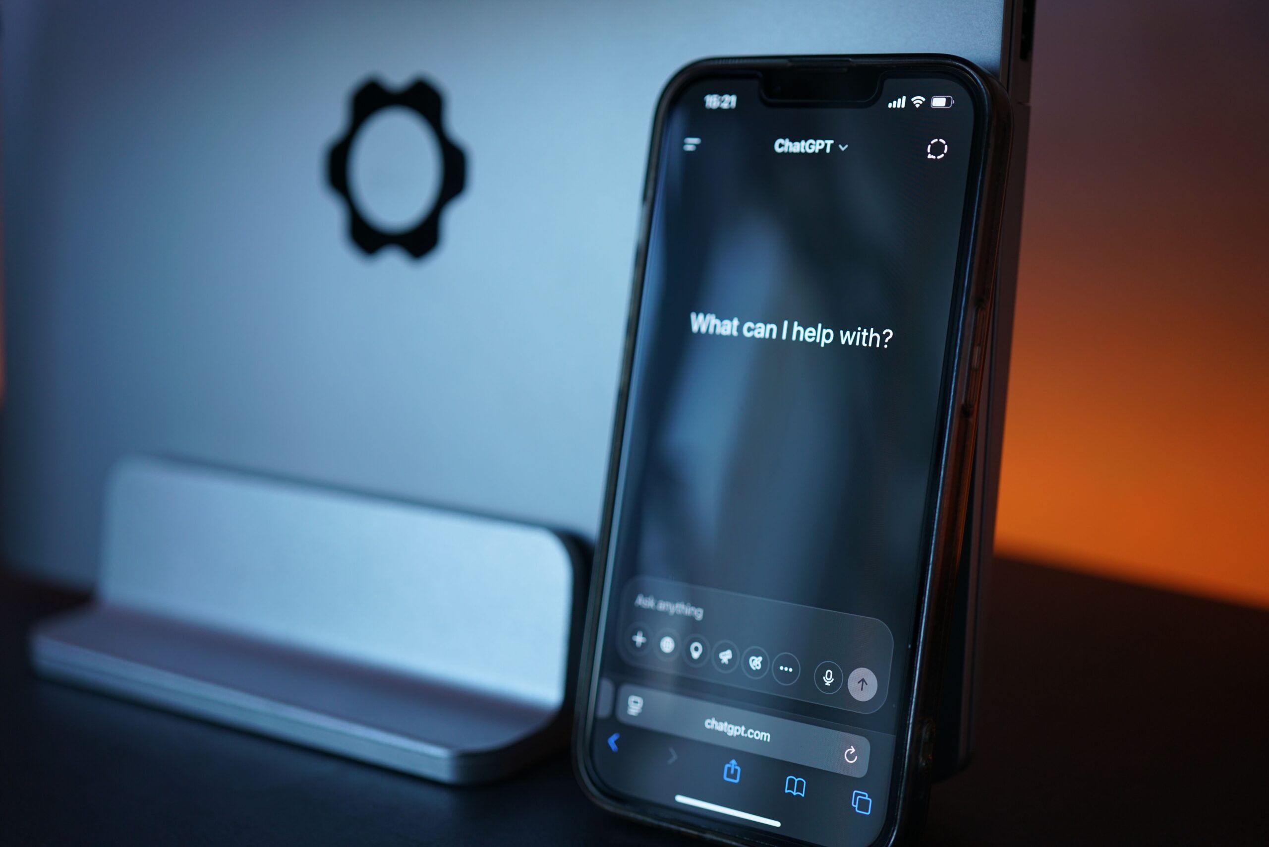 Smartphone displaying ChatGPT beside a laptop in blue and orange lighting.