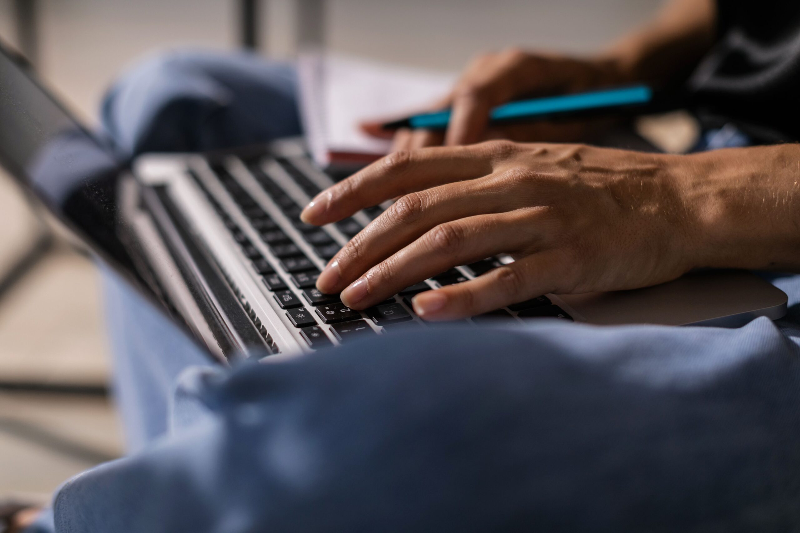 Close-up of a person typing on a laptop while holding a pen and notebook.