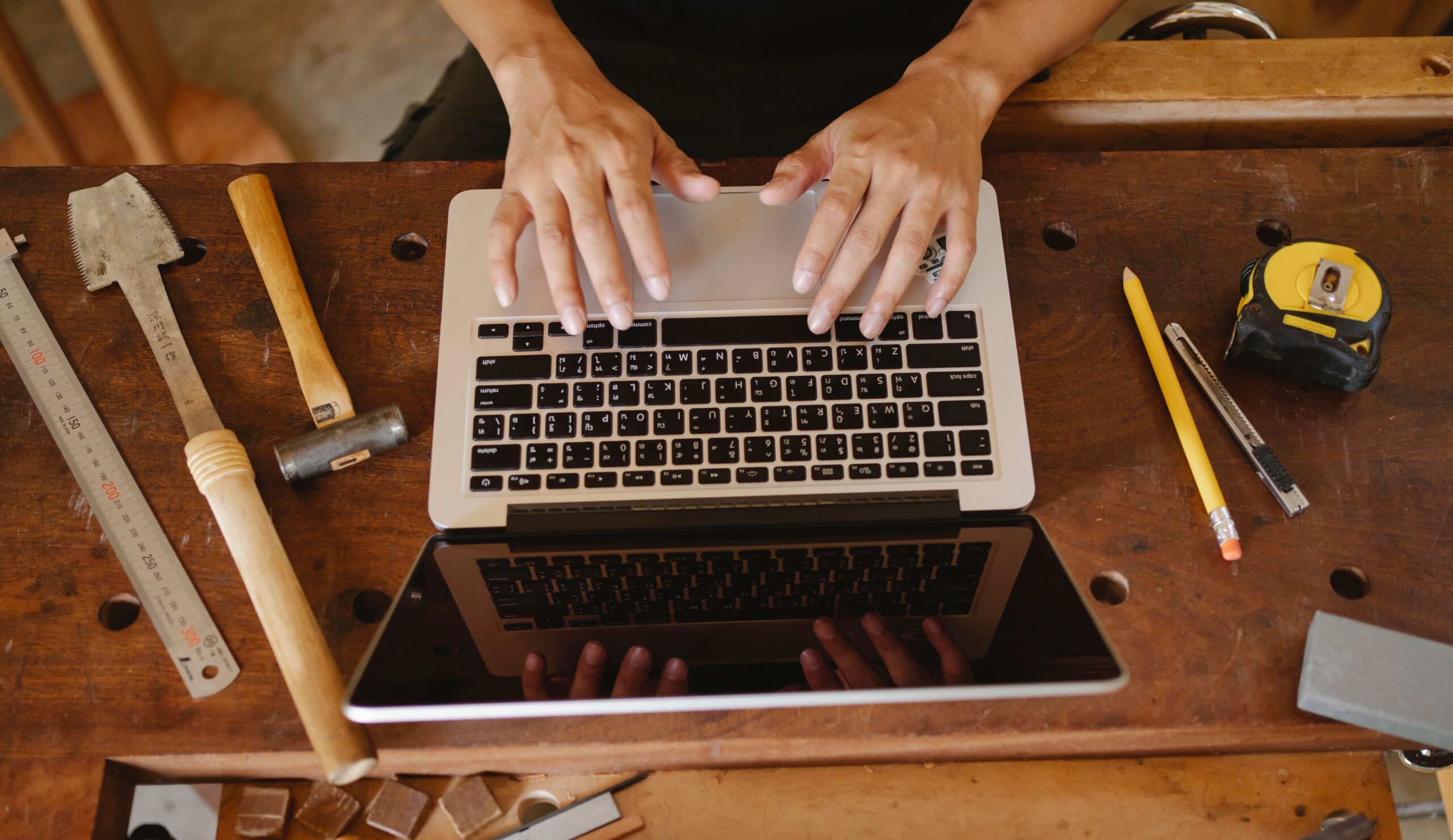 Person using a laptop at a woodworking bench surrounded by measuring and hand tools.