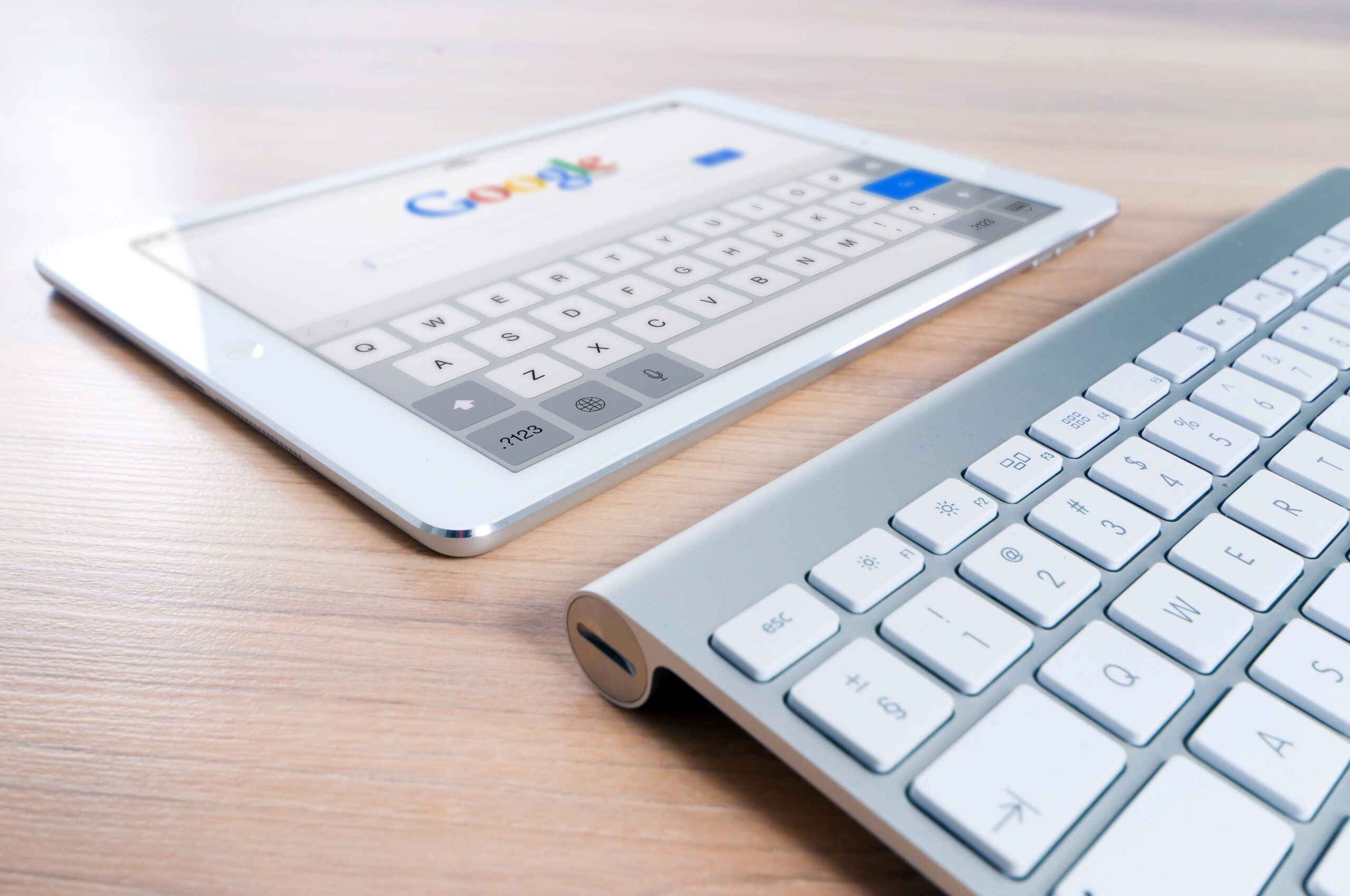 Tablet displaying a Google search page beside a wireless keyboard on a wooden desk.