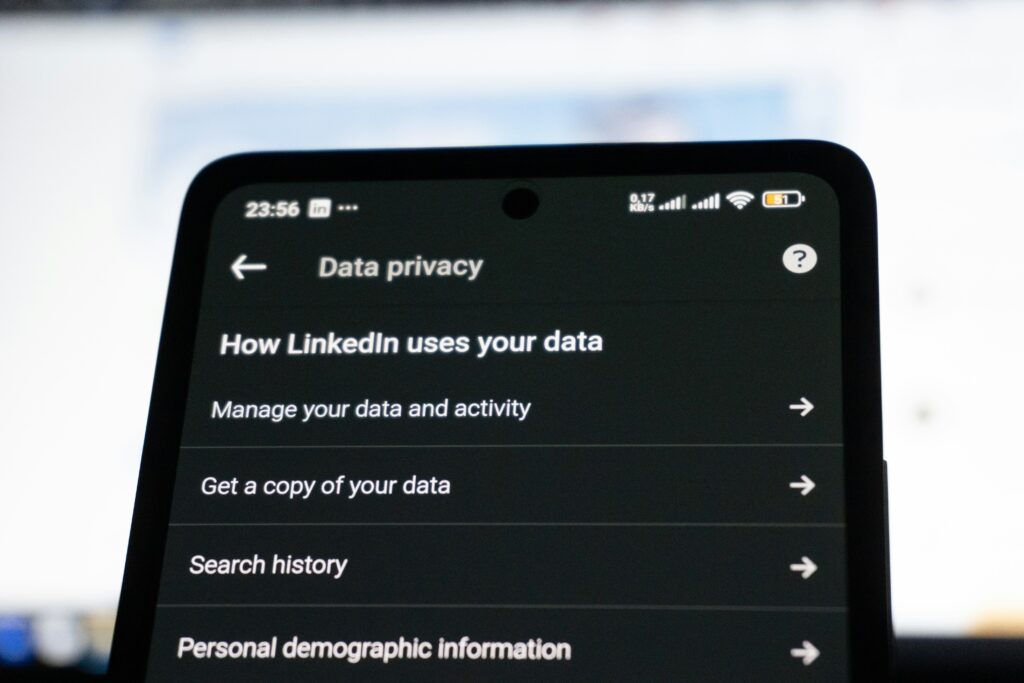 LinkedInのデータプライバシー設定画面をスマートフォンにクローズアップした画像。個人データの管理、アカウント情報のダウンロード、検索履歴の表示などのオプションが含まれている。